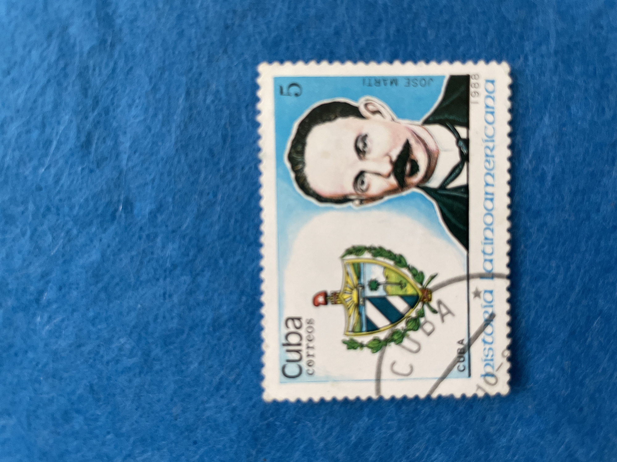 This is a close-up, high-angle shot of a postage stamp placed on a textured blue surface. The stamp is rectangular with perforated edges. The stamp features a portrait of a man with a prominent mustache, facing slightly to the viewer's right. He is depicted in a black coat and tie. To the left of the portrait, a crest or coat of arms is displayed, adorned with green leaves. The background of the stamp is a pale blue sky.
Visible text on the stamp includes:
- "5" in the upper left corner.
- "JOSE MARTI" above the man's head.
- "1988" vertically on the right side.
- "Latinoamericana" vertically on the right side, below "1988".
- "Cuba" and "correos" vertically on the lower left side.
- "CUBA" in smaller text below the crest.
The stamp appears to be in good condition, with vibrant colors. The blue surface it rests on has a fabric-like texture, possibly felt or a similar material. The lighting is even, with no harsh shadows, suggesting a well-lit indoor environment. The overall impression is that of a collectible item being displayed. No specific time of day, weather, or emotional cues are discernible beyond what is depicted on the stamp itself. The location context provided (Perm, Russia) is not directly evident from the image content.