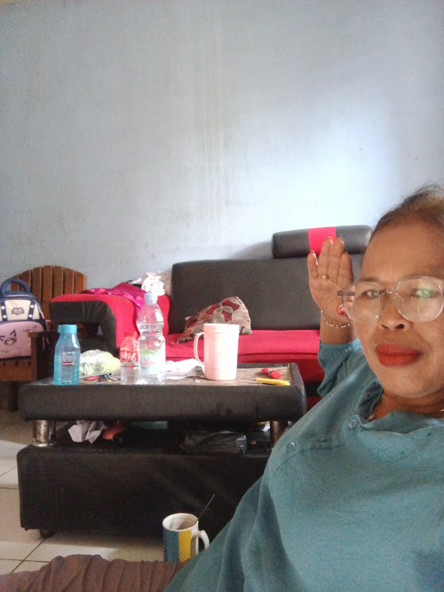 In a room in Warungkiara, Indonesia, a woman with glasses and bright lipstick sits on a couch. She is wearing a teal top and has her right hand raised. In front of her is a black coffee table with several items on it, including two water bottles, a white mug, and a pink cup. Behind the coffee table is a red couch with a black headrest. To the left of the couch is a wooden chair with a backpack resting on it. The room has light blue walls.