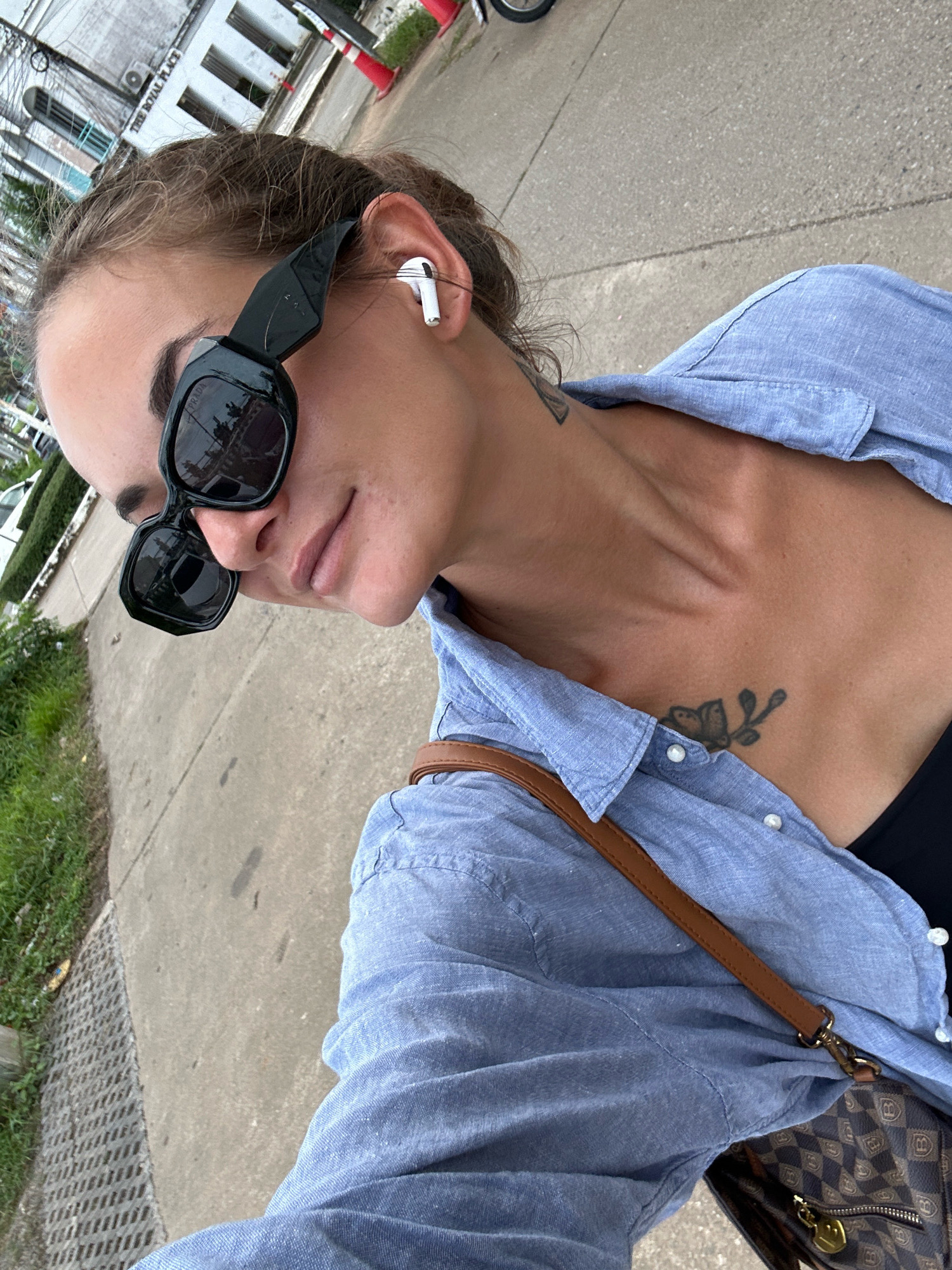 A young woman, wearing large black sunglasses and a light blue button-down shirt, is seen from a close-up, slightly overhead angle. She has white wireless earbuds in her ears and a tattoo on her chest of a butterfly on a branch. Her hair is pulled back, and she is looking slightly away from the camera with a neutral expression. A brown strap from a purse is visible across her shoulder, and the purse itself, with a checkered pattern and prominent "B" logos, is also visible. The background shows a concrete sidewalk and some green grass, with a building and power lines in the distance. The image appears to have been taken outdoors during the daytime, possibly in an urban or suburban setting in Kathu, Thailand, given the background elements and architecture.