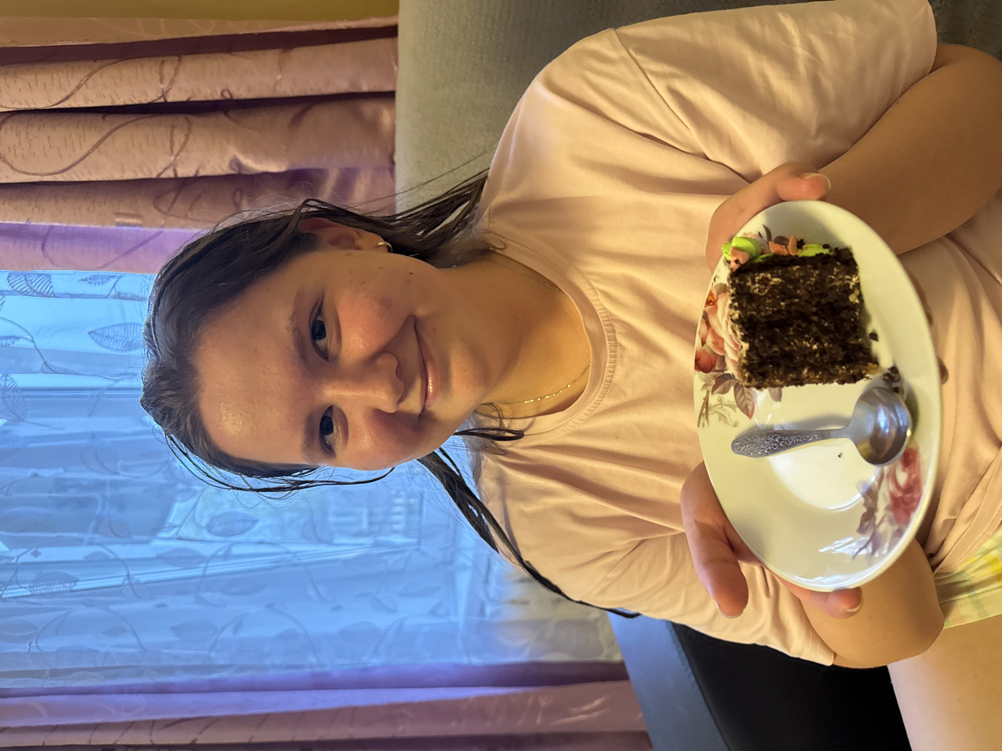 A young woman holds a piece of chocolate cake on a floral plate with a spoon. She is wearing a pink t-shirt and a gold necklace. The background shows a curtained window and a gray sofa. The photo was taken indoors. There is no indication in the image that would allow it to be geo-located specifically within Бородулино, Russia.