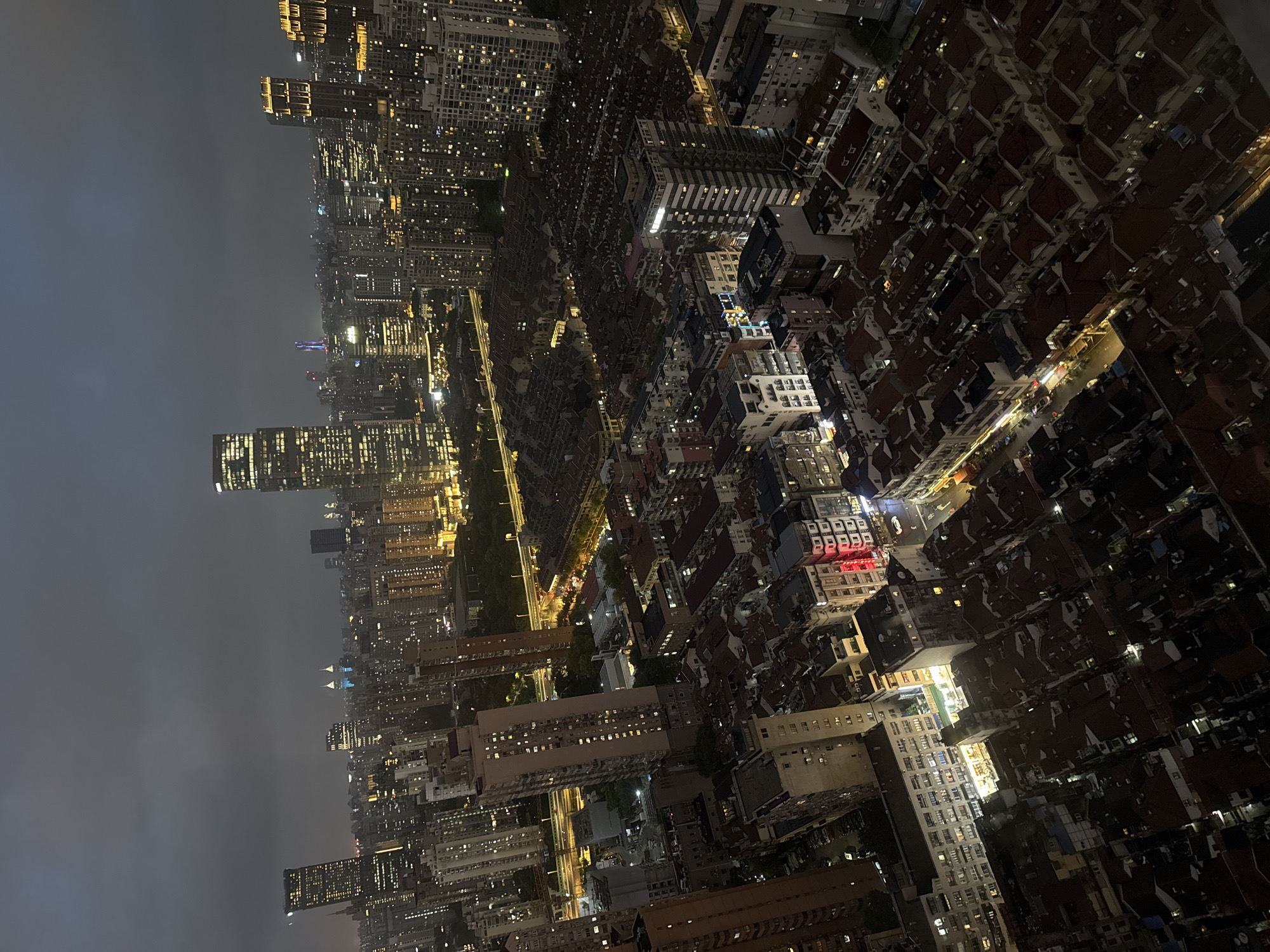 This is a night aerial view of the city of Shanghai, China, likely from a high-rise building. The scene is dominated by a dense urban landscape with a mix of modern skyscrapers and older, low-rise buildings with reddish-brown roofs. Many buildings are illuminated, with windows lit up and some larger structures featuring architectural lighting.
In the foreground and middle ground, there are many densely packed, low-rise residential or commercial buildings with a consistent architectural style, featuring dormer windows and tiled roofs. Streets with vehicles and streetlights are visible between these buildings. A multi-lane elevated road or highway cuts through the mid-ground, also illuminated by streetlights.
In the background, towering skyscrapers rise into the dark, overcast sky. Some of these skyscrapers are very tall and slender, with illuminated facades and windows. One particularly tall, slender building on the far left has many horizontal bands of light. In the distance, several prominent skyscrapers stand out, including one with distinctive pointed tops that are illuminated in blue. Other buildings in the background are lit with warm yellow and white lights, creating a city-wide glow against the twilight sky. The overall impression is of a sprawling, vibrant metropolis at night. There are no visible people or discernible actions taking place.