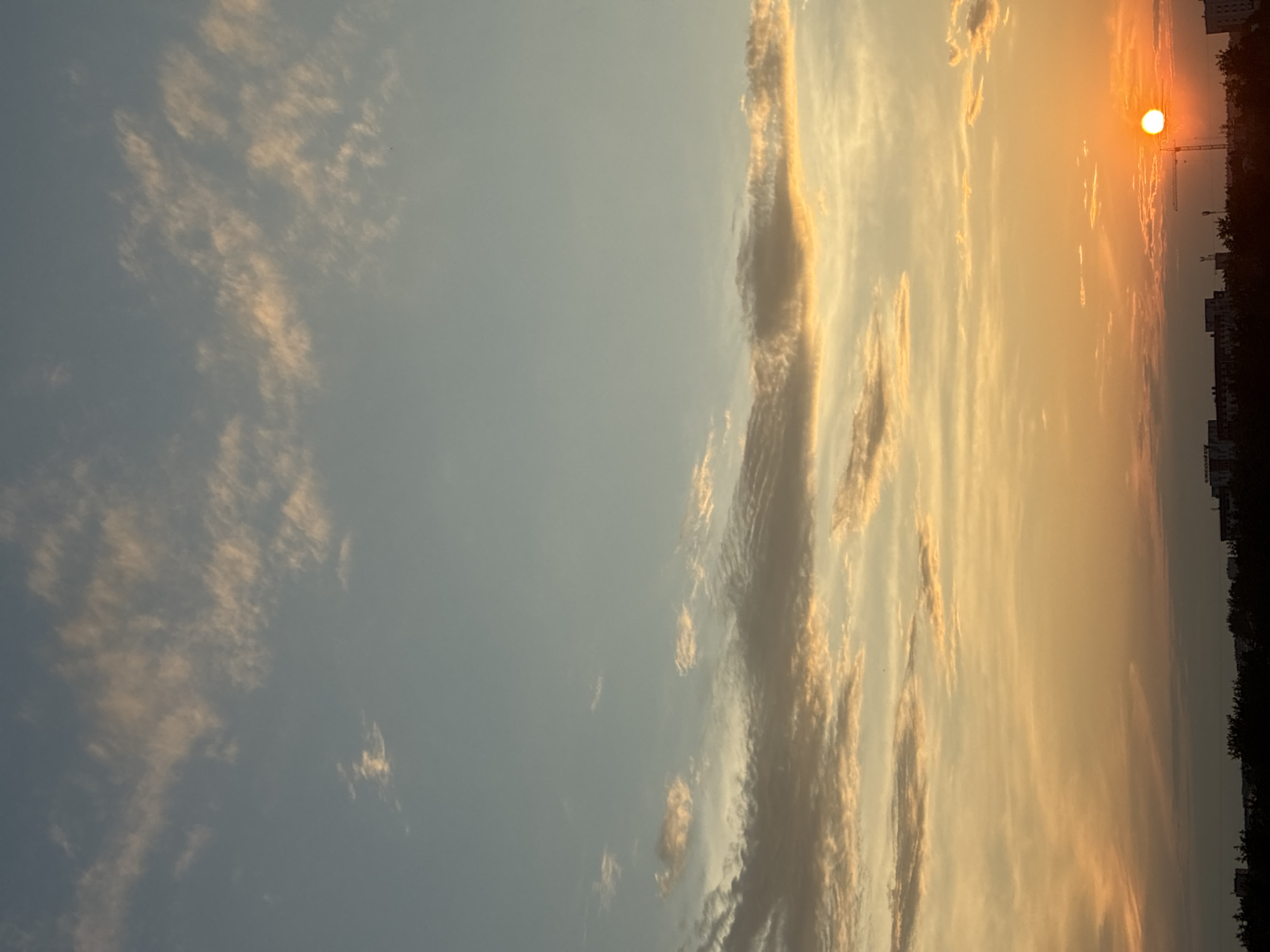 A wide shot captures a vibrant sunset over the city of Unknown, Unknown. The sky is a canvas of warm hues, transitioning from a soft blue at the top to a fiery orange and yellow near the horizon. Wispy clouds are scattered across the sky, catching the sunlight and creating streaks of pink and gold. The sun, a brilliant orb, is partially obscured by the horizon, casting a warm glow. Below the sky, a dark silhouette of buildings and trees forms the city's skyline. A construction crane is visible among the buildings, suggesting ongoing development. The overall scene is one of natural beauty and urban life coexisting.