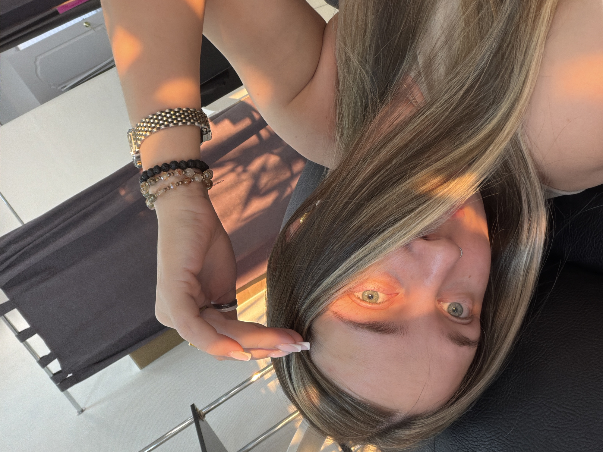 A selfie taken by a blonde woman indoors, likely in Chaikovsky, Russia. She has fair skin, light eyes, a nose piercing, and is wearing a few bracelets and a ring. The lighting suggests it's sunset. A black object and a gray folding chair are visible in the background.