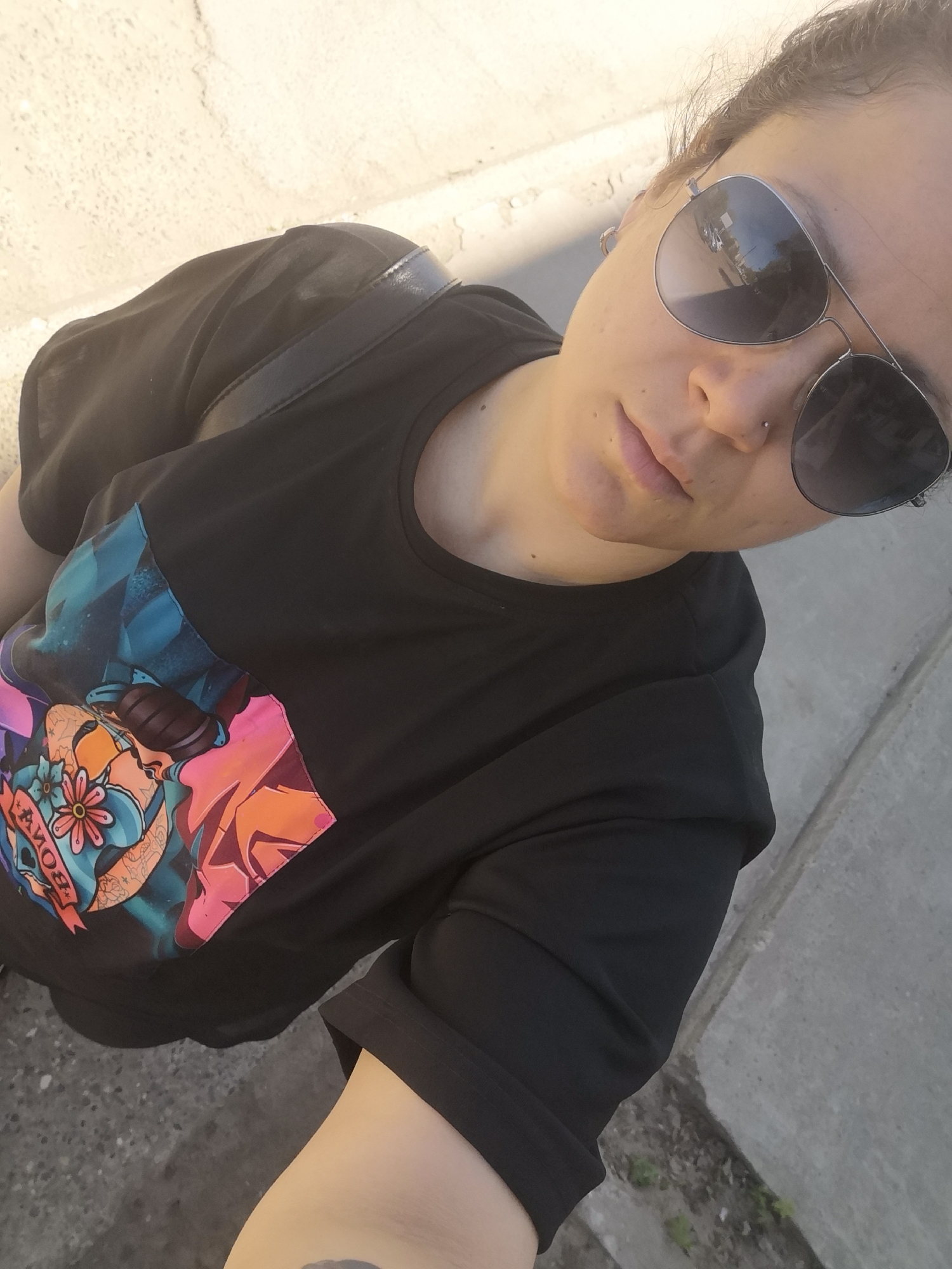 A woman with sunglasses is posing for a selfie outdoors. She is wearing a black t-shirt with a colorful graphic design. She has a nose ring and earrings. A black bag strap is visible on her shoulder. The background shows a concrete sidewalk and curb. It is not possible to confirm the location is in Chaikovsky, Russia from the image alone.