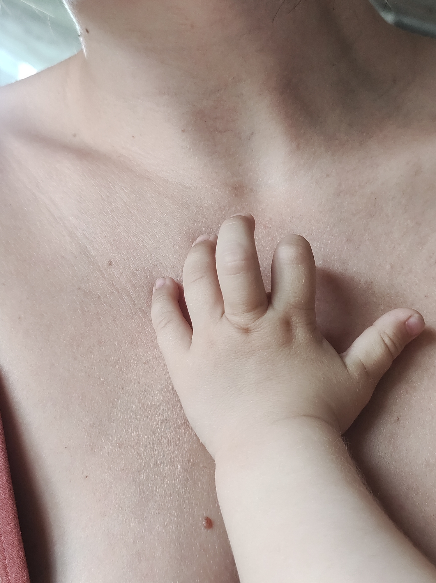 The image shows a close-up of a person's chest with a child's hand resting on it. The skin is fair and the hand is small and chubby. There are a few moles on the chest. The image is neutral in tone and does not convey any specific emotion or activity. The location is not determinable from the image content.