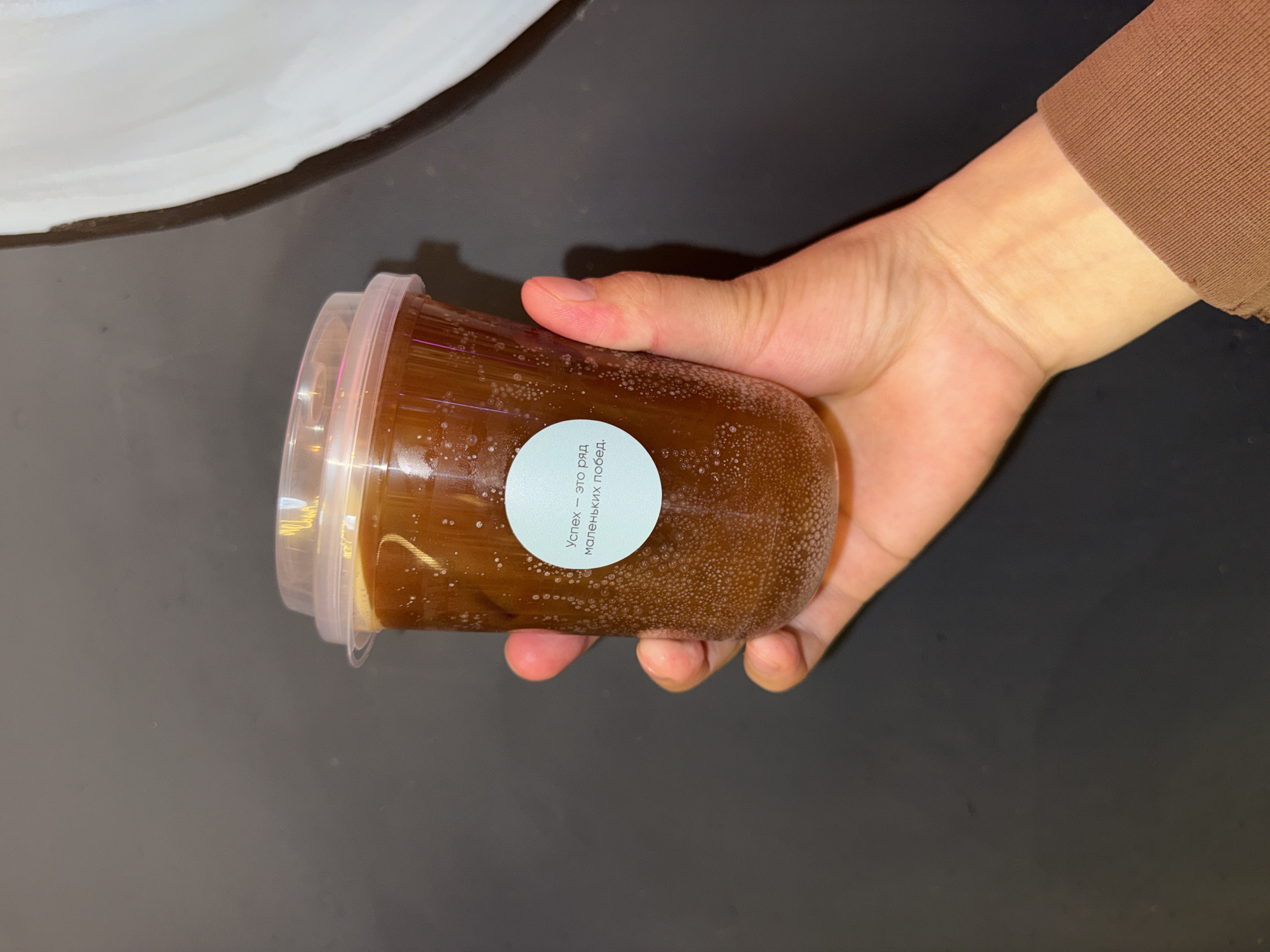 A hand holds a transparent plastic cup filled with a dark brown beverage, likely coffee or iced tea, with visible bubbles. A small, round, light blue sticker with white text in Russian is affixed to the cup. The text translates to "Success is a series of small victories." The background is dark gray. The image is a close-up shot, suggesting it was taken indoors, possibly in a home or cafe environment. The presence of the cup and hand indicates a person is interacting with the beverage. The location in Veshki, Russia, is not discernible from the visual elements of the image itself.
