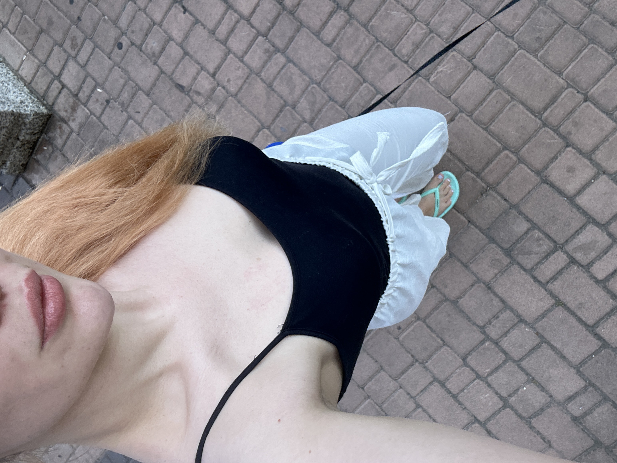 The image is an overhead shot of a woman from the shoulders down, standing on a paved sidewalk in Odesa, Ukraine. She has light skin and reddish-blonde hair. She is wearing a black spaghetti strap tank top and loose, white, flowy pants tied at the waist with a drawstring. She is also wearing light blue flip-flops. The sidewalk is made of reddish-brown interlocking pavers. In the background, a portion of a stone structure is visible, as well as what appears to be a dark leash extending out of the frame to the right. The scene appears to be outdoors, possibly in a public area like a park or street.