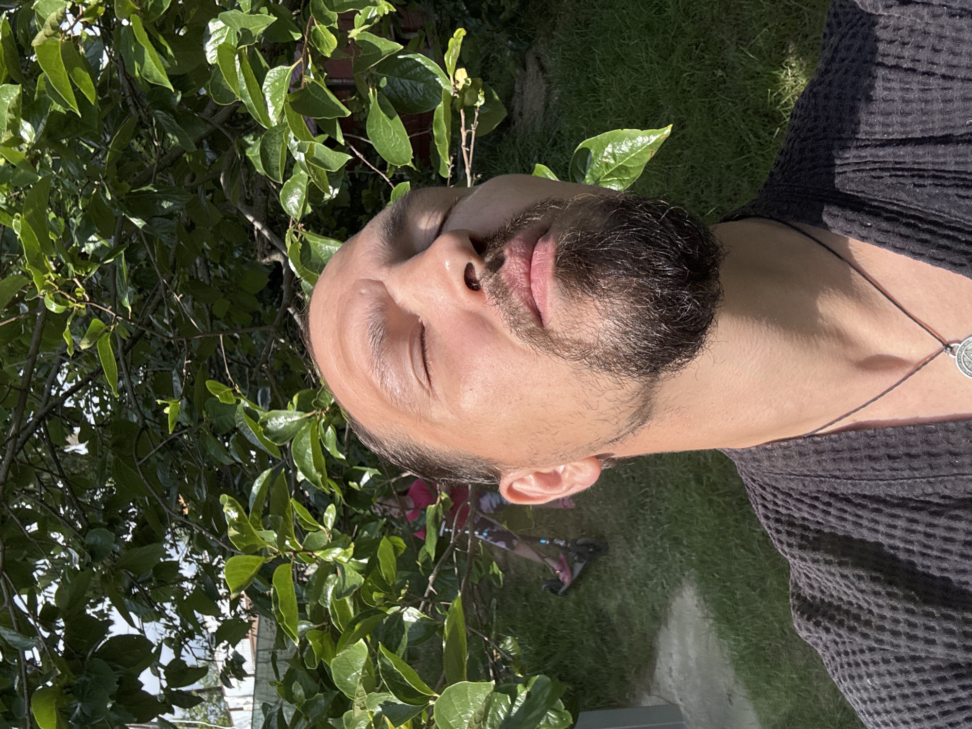 The image is a close-up, Dutch angle shot of a person's face tilted upwards, with their eyes closed. The person has light skin, a beard, and dark hair. They are wearing a dark, textured robe or shirt and a necklace with a round pendant. The background features lush green foliage and grass, suggesting an outdoor setting. Sunlight filters through the leaves, creating dappled shadows and highlights on the person's face and the surrounding environment. The overall mood appears serene and relaxed, possibly indicating the person is enjoying the sun or meditating. The time of day is likely daytime, given the bright sunlight. The context provided indicates the location is Sochi, Russia, which is known for its warm climate and abundant greenery, consistent with the visual elements. No text is visible in the image.