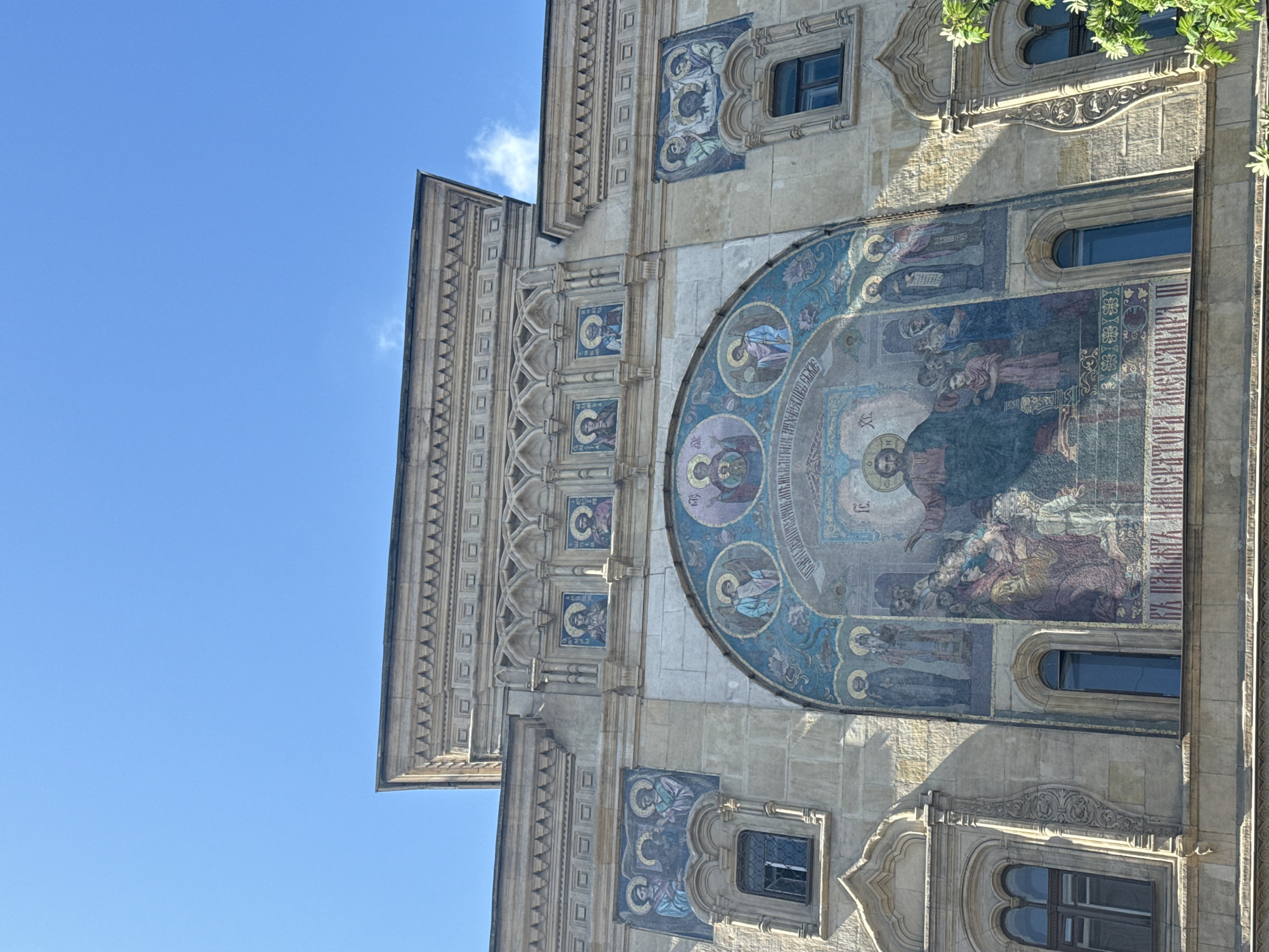 A low-angle, rotated view shows the exterior of a building, likely a church, in Saint Petersburg, Russia. The structure is made of light-colored stone or concrete and features intricate carvings and architectural details. A large, circular mosaic dominates the facade, depicting religious figures in a traditional Orthodox Christian style. Smaller mosaic panels with portraits adorn the walls. The building also has several arched windows. The sky above is clear and blue, with a few wisps of clouds. Green foliage can be seen at the top of the frame.