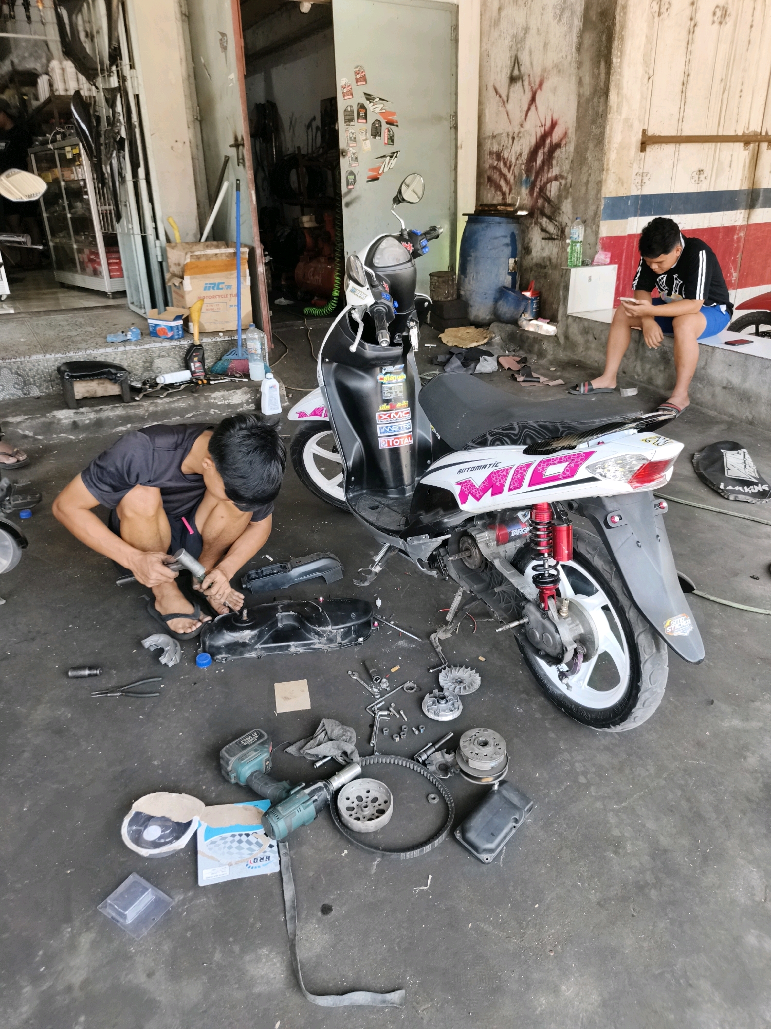 In Silian Dua, Indonesia, a man is working on a scooter in a cluttered workshop. The man, wearing a black t-shirt and shorts, is crouched down and appears to be using a tool on the scooter's engine components which are spread out on the concrete floor. Nearby, another person is seated, looking at their phone. The workshop is filled with tools, parts, and various items, suggesting an active repair space. A blue barrel and a blue broom are visible in the background, along with a wall covered in graffiti and posters.