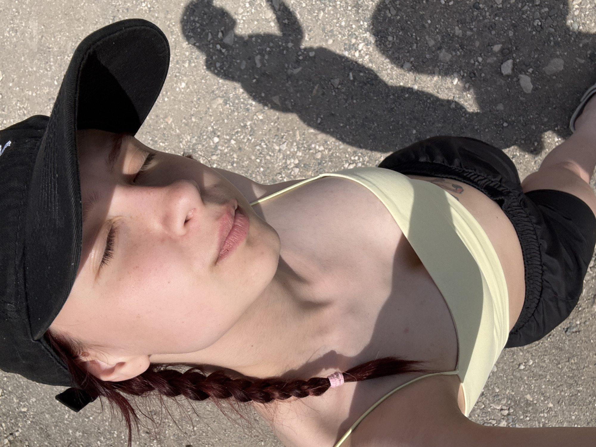 A person is seen from an overhead perspective on a sunlit, possibly concrete, outdoor surface. The person is wearing a black baseball cap, a pale yellow spaghetti strap top, and black athletic shorts. A dark braid with a pink band at the end is visible. A tattoo is noticeable on the lower torso above the shorts. The shadow of a second person is cast on the ground, partially obscured by the body of the person being photographed. The environment suggests a location in Khvalynsk, Russia. The exact location within the city is unknown.