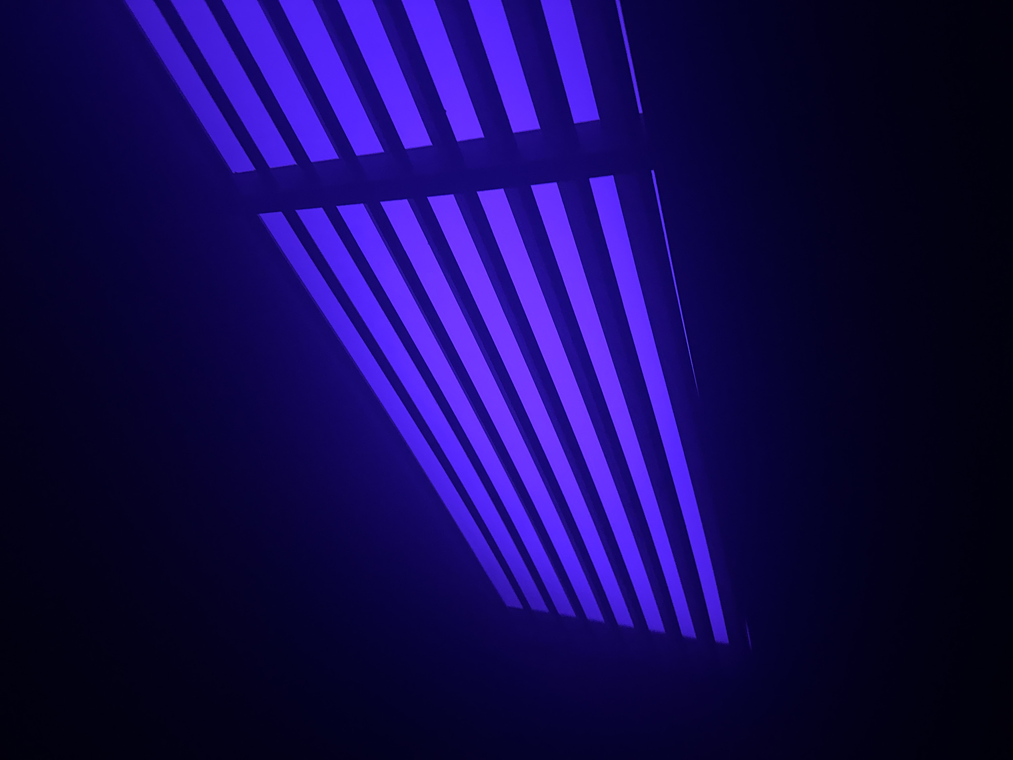 The image depicts a blue, striped pattern. The pattern appears to be formed by the reflection of light through a set of parallel bars or slats. The background is dark, with a gradient transitioning from a lighter blue near the striped pattern to a deeper black further away. There are no visible people, subjects, actions, or interactions in the image. The image does not provide enough information to determine a specific location in Paris, France.