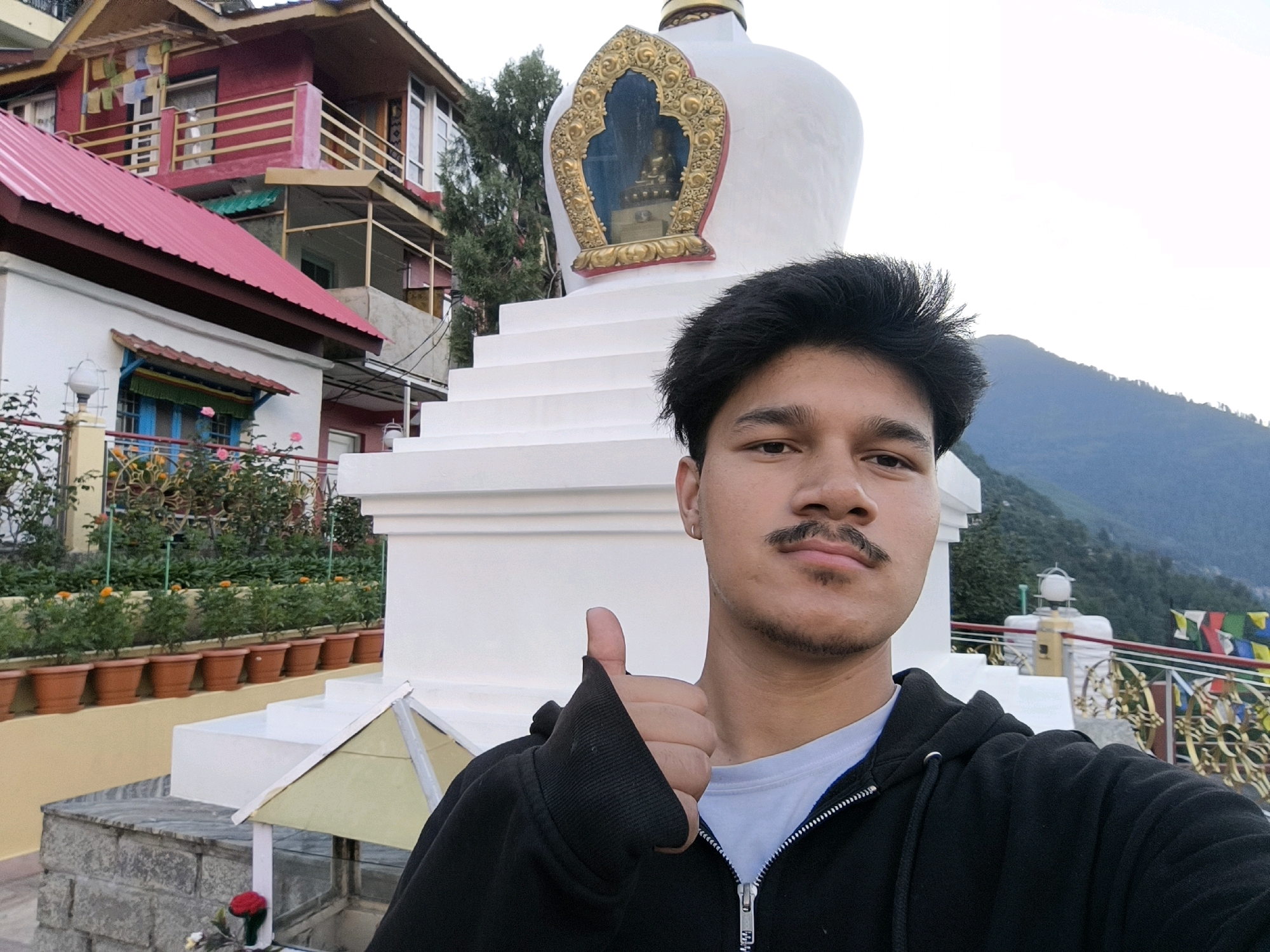 A young man with dark hair and a mustache is taking a selfie in front of a white stupa. He is wearing a black hooded sweatshirt and a light blue t-shirt. His left arm is raised, and he is giving a thumbs-up. The stupa has a golden decorative frame with a Buddha statue inside. Behind the stupa, there are mountains and prayer flags. To the left of the stupa, there is a red building with balconies and windows, and potted plants are visible on a ledge. The image appears to be taken outdoors during daylight. The location is identified as Kalath, India.