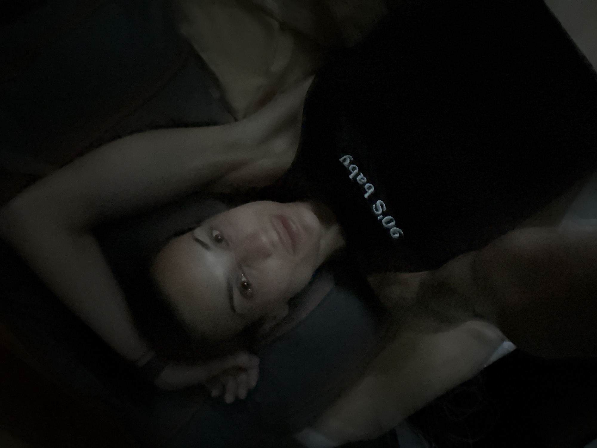 A low-angle, dimly lit, indoor selfie shows a woman lying down with her head slightly tilted and her right arm bent behind her head. She is wearing a black tank top with the words "90's baby" embroidered in light blue text. Her left arm is extended downwards and out of view. The background is dark and appears to be a bed or couch. The image is grainy and out of focus in some areas. There are no identifiable landmarks or context that would suggest a location in Pleven, Bulgaria.