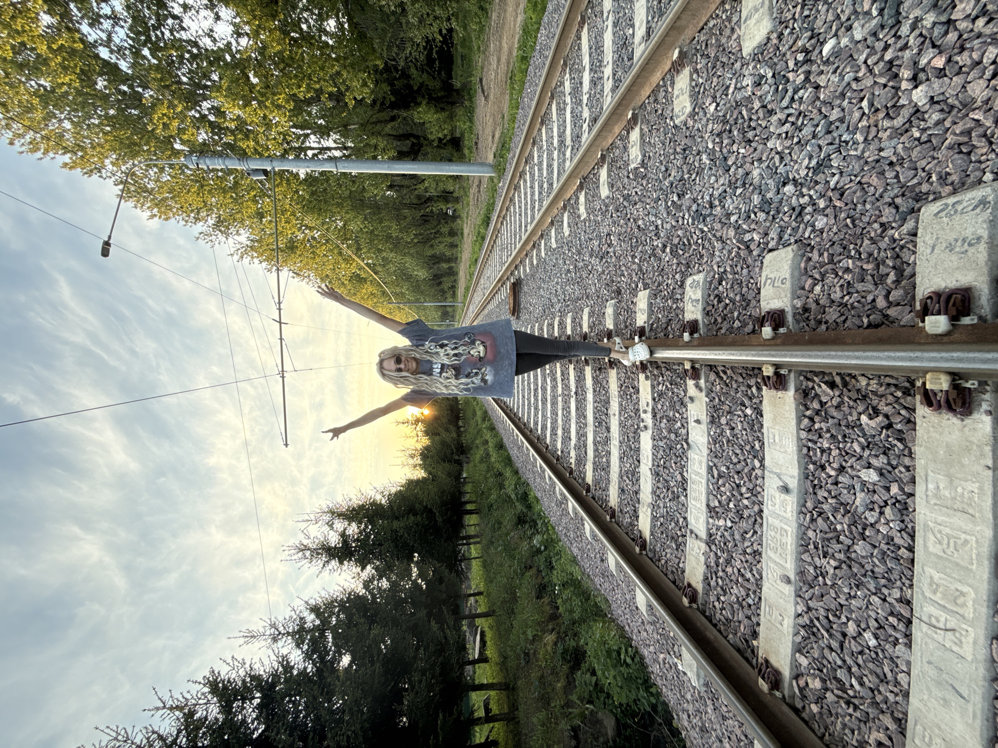 A woman stands on a railway track. The tracks are lined with gravel and concrete sleepers. Overhead power lines are present. Trees and sky are visible in the background, along with a sunset. The location is Заволжье, Russia.