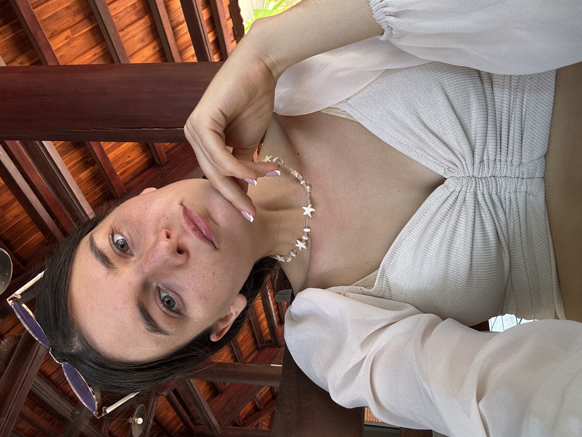 A woman with fair skin, dark hair, and light-colored eyes is seen in a selfie-style image taken from a low angle. She is wearing a light-colored top with sleeves that gather at the cuffs and a necklace with starfish-shaped beads. The woman is touching her face with her hand, and a pair of sunglasses are partially visible near her hair. Above her is a wooden structure with a reddish-brown hue, possibly part of a building or gazebo. Light is filtering through the structure, suggesting an outdoor setting. Given the context of Mũi Né, Vietnam, the location is likely a resort or outdoor seating area with a traditional Vietnamese-style wooden roof structure. The image does not provide sufficient detail to pinpoint the exact location in Mũi Né.