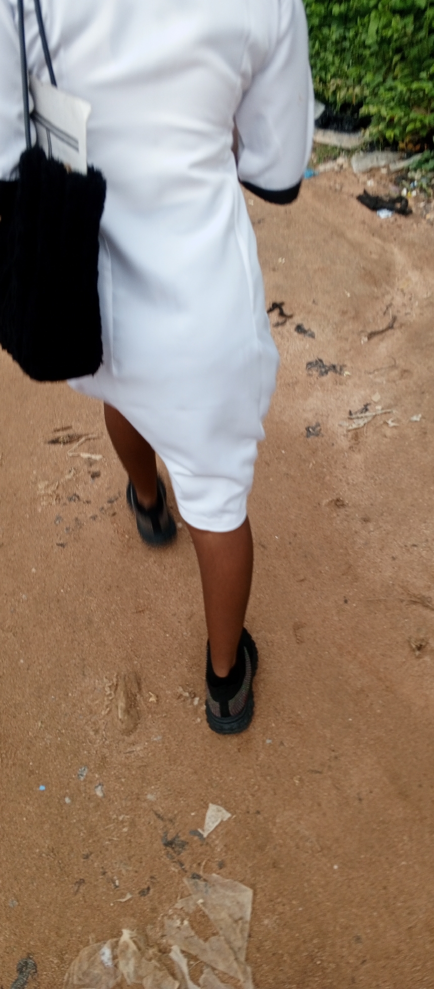 A person walks on a sandy path in Kuson, Nigeria. They are wearing a white robe with black trim and black athletic shoes. A black fuzzy bag hangs from their left shoulder. The path is made of dirt and sand, with scattered debris including plastic and plant matter. In the background, green foliage is visible.