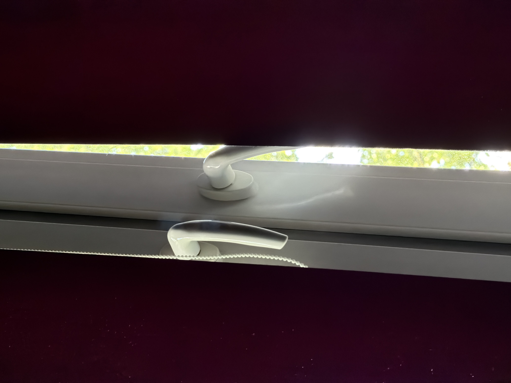 The image shows a close-up view of two white window handles on a white window frame. The window is set against a dark purple background, possibly a wall or curtain. Sunlight is streaming through a narrow gap in the window, illuminating the handles and casting reflections. The scene appears to be an interior shot, with the window likely providing a view of the outdoors, as indicated by the bright green light filtering through the gap. The image does not provide any information about the city of Khimki, Russia.