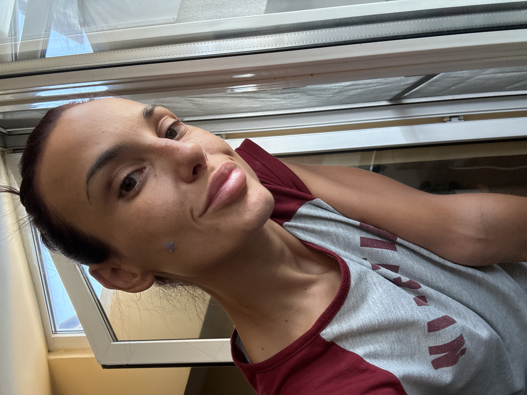 A woman is taking a selfie near a window in Pleven, Bulgaria. She is wearing a grey and maroon t-shirt with a graphic design. She has a light purple star sticker on her cheek. The window has a screen and is partially open. Sunlight is coming through the window and reflecting off the glass. The background shows a wall and the sky.