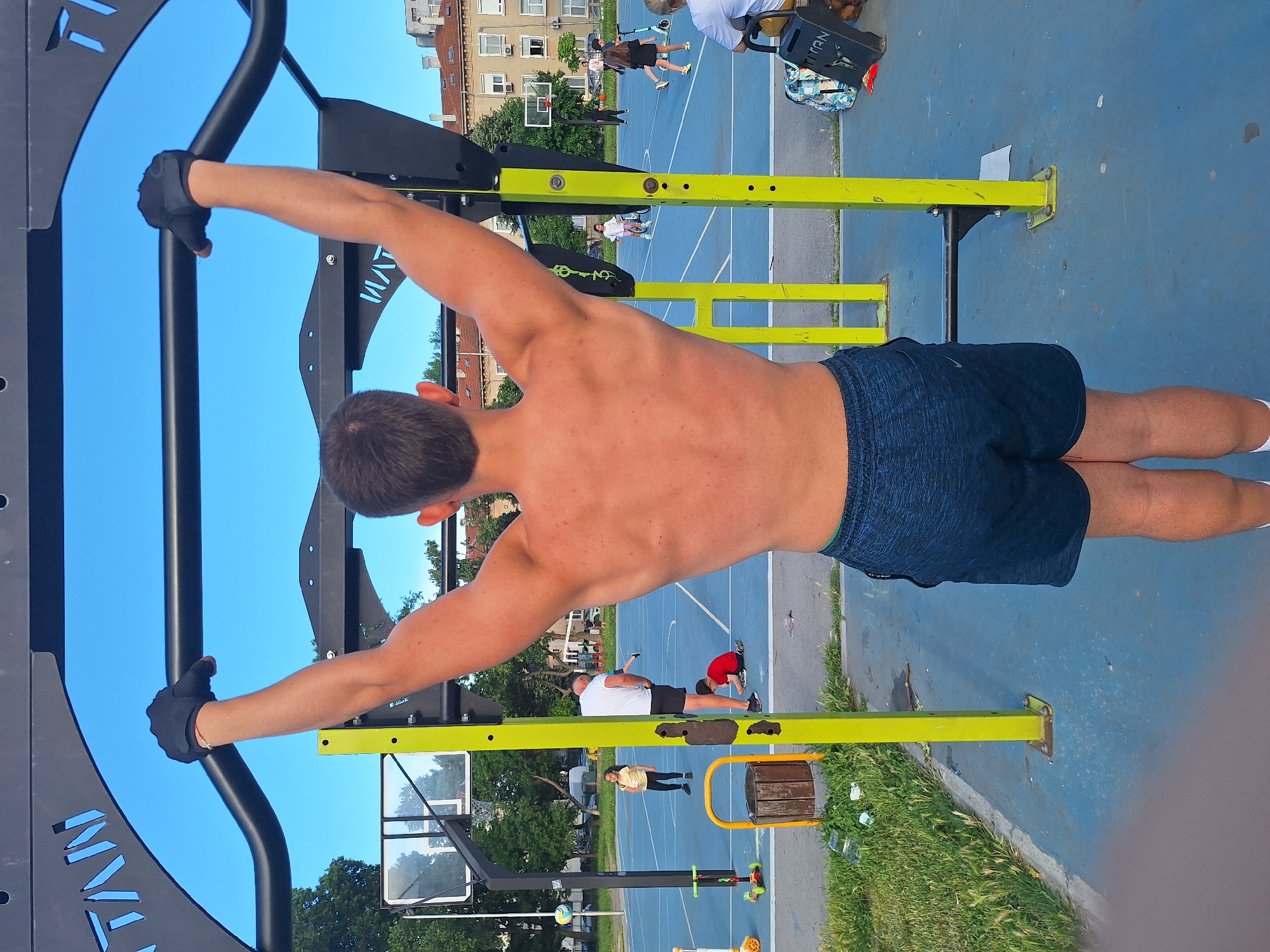 The image depicts a young man performing a pull-up on an outdoor fitness apparatus in Varna, Bulgaria. The man is viewed from behind, with his arms fully extended as he grips the horizontal bar. He is wearing black fingerless gloves and dark blue shorts. The fitness equipment is painted bright yellow and black, with the brand name "TITAN" visible on the frame.
The setting appears to be a public park or recreational area, evidenced by a blue basketball court in the background with several children playing. There are also trees, buildings with terracotta roofs, and streetlights visible in the distance. The sky is clear and blue, indicating fair weather. A wooden trash bin and some scattered debris are present on the ground near the fitness equipment. The overall scene suggests a sunny day in a vibrant urban outdoor space.