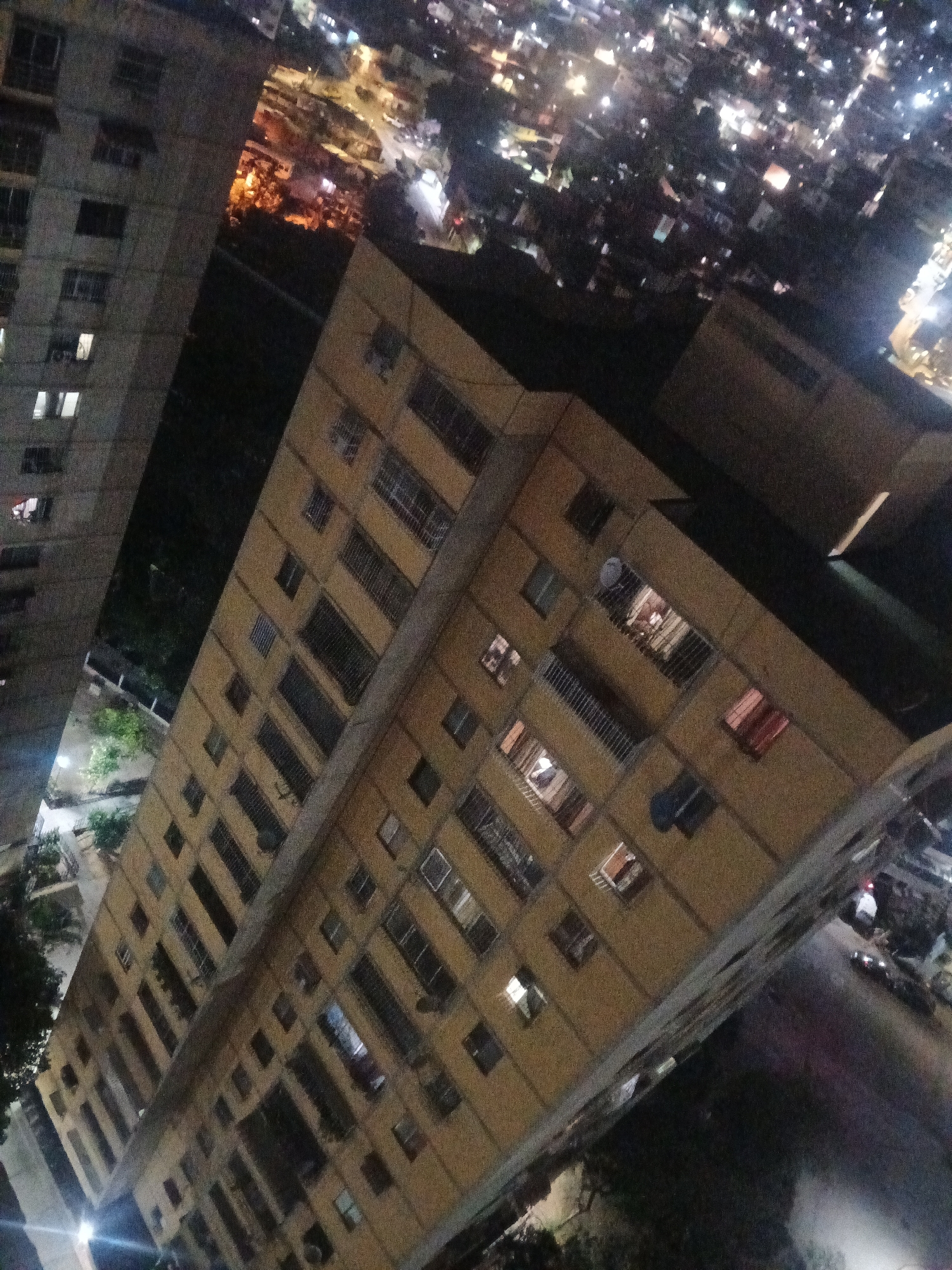 The image depicts a high-angle view of residential buildings at night. The buildings are illuminated by internal lights, with some windows showing activity. The city's lights are visible in the background, suggesting an urban setting. The scene appears to be located in Caracas, Venezuela, given the architectural style and the general urban layout. The absence of distinct landmarks makes precise localization within Caracas challenging. The image provides limited detail, focusing on the buildings and the nightscape. No people are visibly identifiable in the scene. The overall environment is urban and residential, captured under low-light conditions.