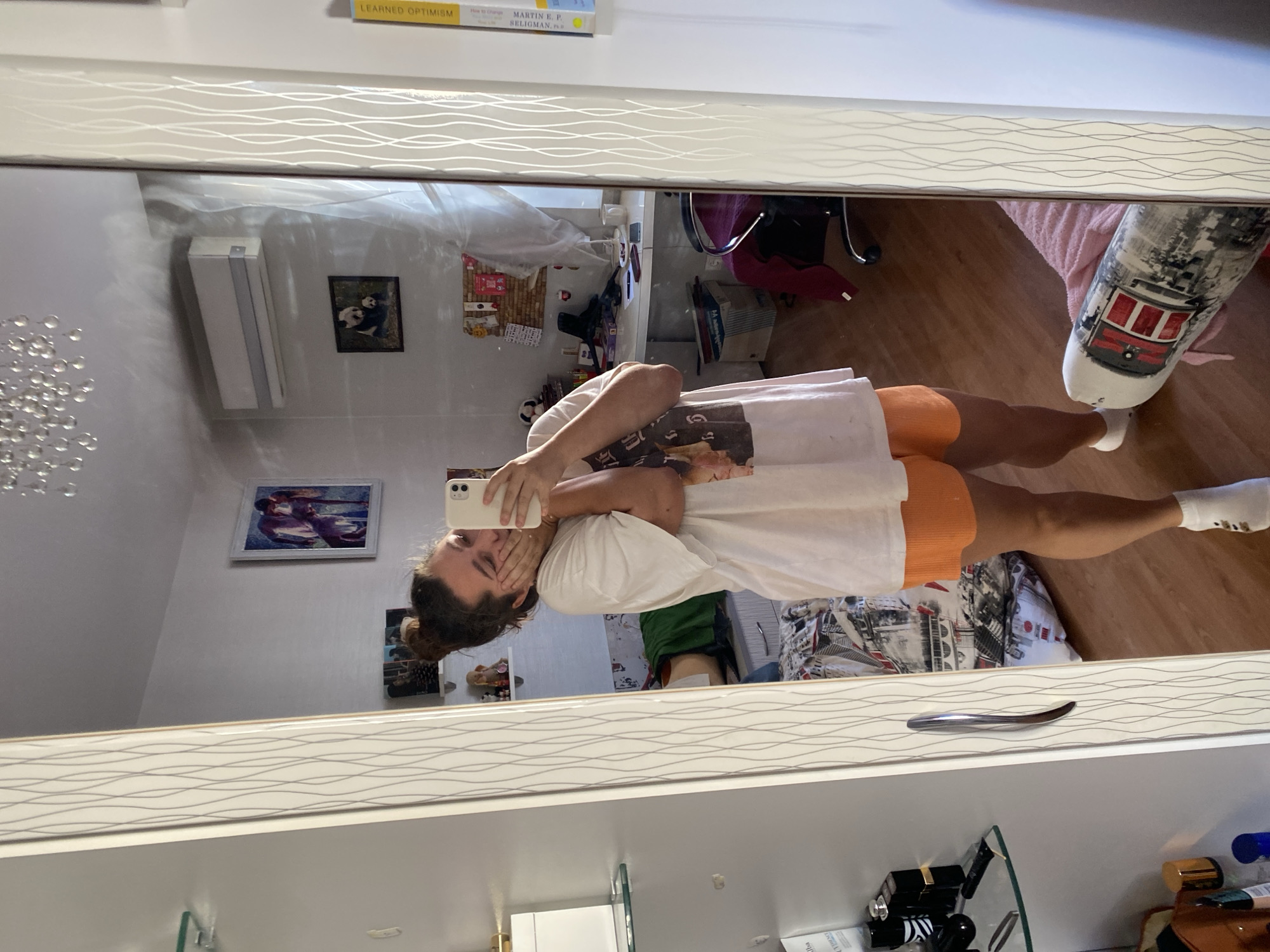 A young woman with her hair in a bun, wearing a white t-shirt and orange shorts, takes a mirror selfie in a bedroom. She is holding her phone in her right hand and resting her left hand on her cheek. The mirror reflects a room with a bed, a desk with a chair, a bookshelf, and a window. On the wall above the desk, there is a framed picture of two pandas and an air conditioning unit. To the left of the woman, a shelf is visible with several bottles of perfume and skincare products. The overall scene suggests a casual and personal moment captured in a domestic setting. The location in the city of Perm, Russia is not ascertainable from the visual information provided in the image.