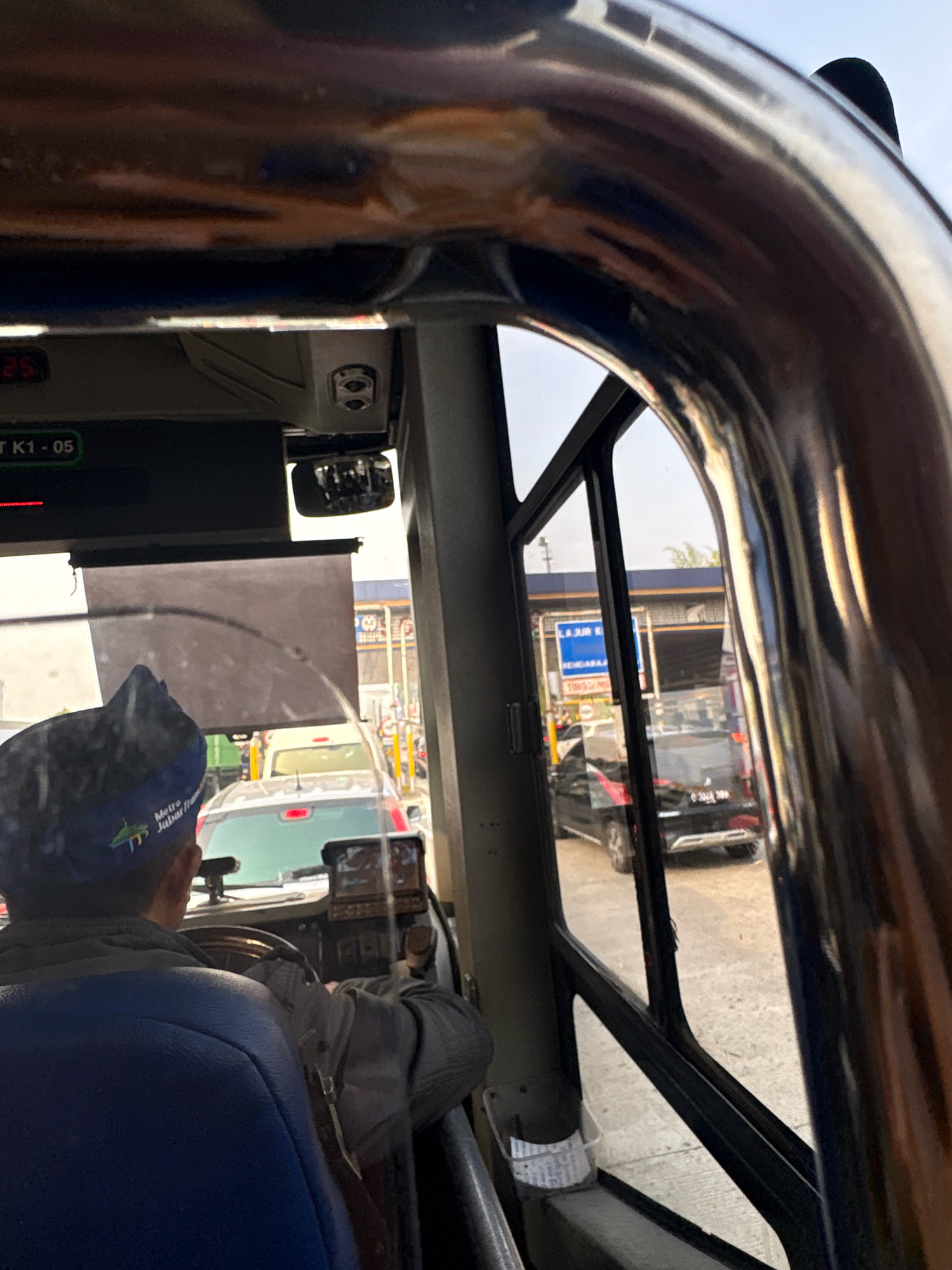 The image depicts the interior of a bus, viewed from the driver's perspective. A person wearing a blue cap with the text "Metro Jawa Barat" is visible driving the bus. Outside the bus, a traffic scene is visible. Cars are lined up at what appears to be a toll booth or a checkpoint. The environment suggests a city street in Parungserab, Indonesia, given the visual cues of traffic and potential infrastructure. The overall lighting suggests it is daytime, with sunlight reflecting off the metallic structures within the bus.