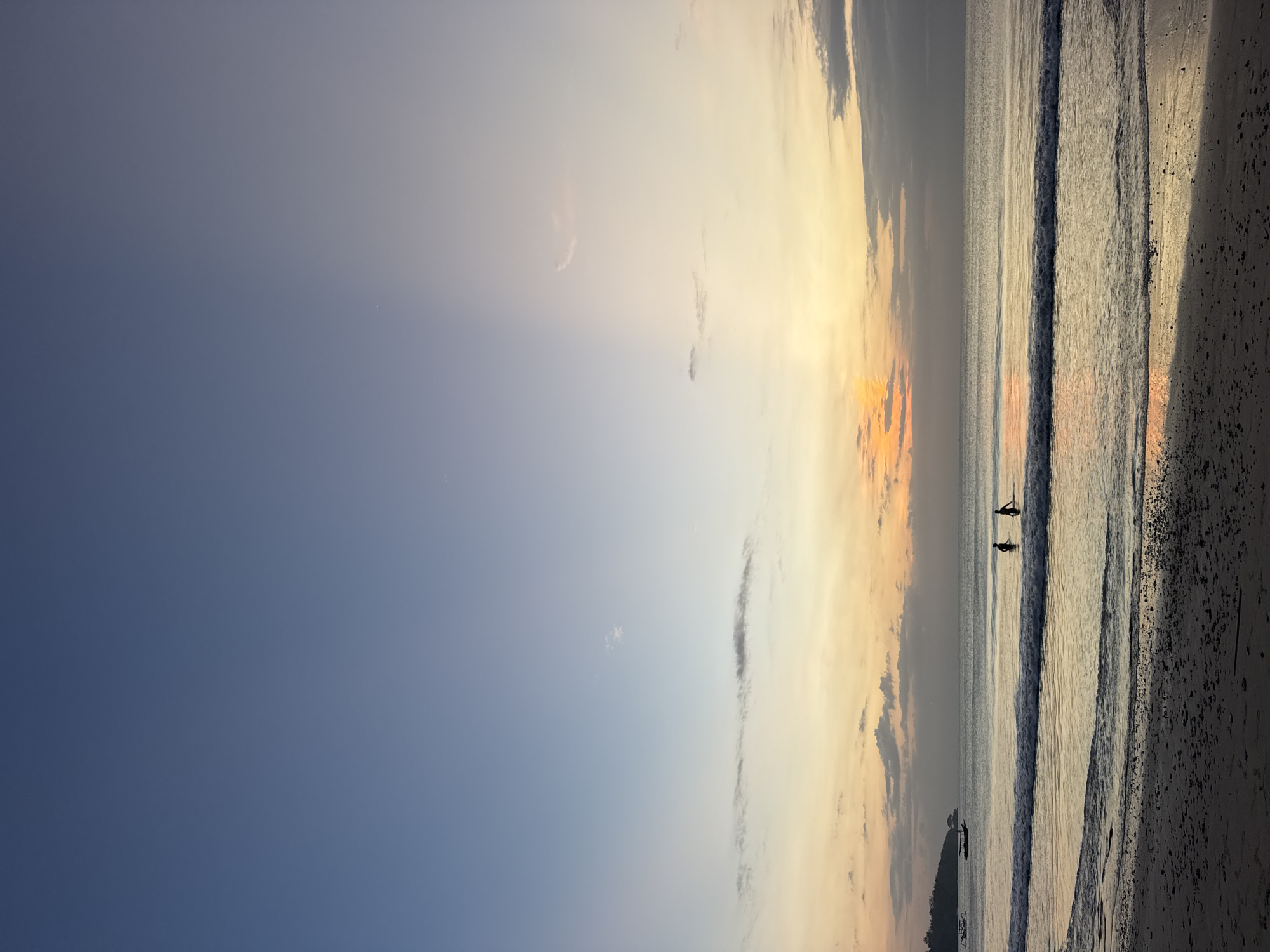 This is a full-frame, vertical, low-angle shot of Kuta Selatan, Indonesia, taken at sunset. The scene is dominated by the vast sky, which transitions from a deep blue at the top to a pale yellow and orange near the horizon. A distinct ray of sunlight cuts vertically through the sky from the upper right, creating a dramatic contrast. The ocean is calm, with gentle waves lapping at the sandy shore. Two silhouetted figures, appearing to be two people, stand in the shallow water, facing the sunset. In the background, a lush green landmass is visible to the left, with a traditional Indonesian boat anchored offshore. The sand in the foreground is dark and textured, with scattered pebbles and footprints.