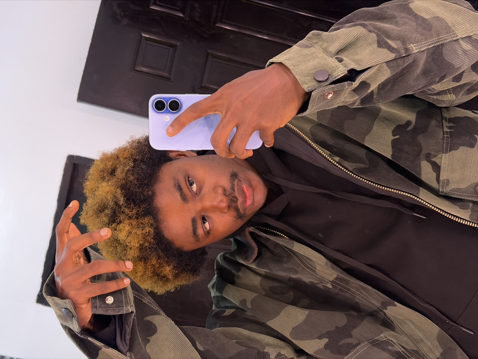 A male individual is depicted in a mirror selfie, located indoors in Igboefon, Nigeria. He has a dark complexion, a mustache, and a short, afro-textured hairstyle with prominent light brown or blonde highlights. He is wearing a black hooded sweatshirt beneath an open, corduroy jacket featuring a green, brown, and black camouflage pattern, equipped with a zipper and snap closures on the cuffs. The subject is holding a light blue or lavender-colored smartphone with a dual-camera system in his right hand, positioned to capture the image. His left hand is raised with fingers splayed, visible above his head in the reflection. The background behind the subject's reflection shows a plain white wall to the left and a dark, paneled door to the right. The subject's gaze is directed slightly to his left, away from the phone's lens.