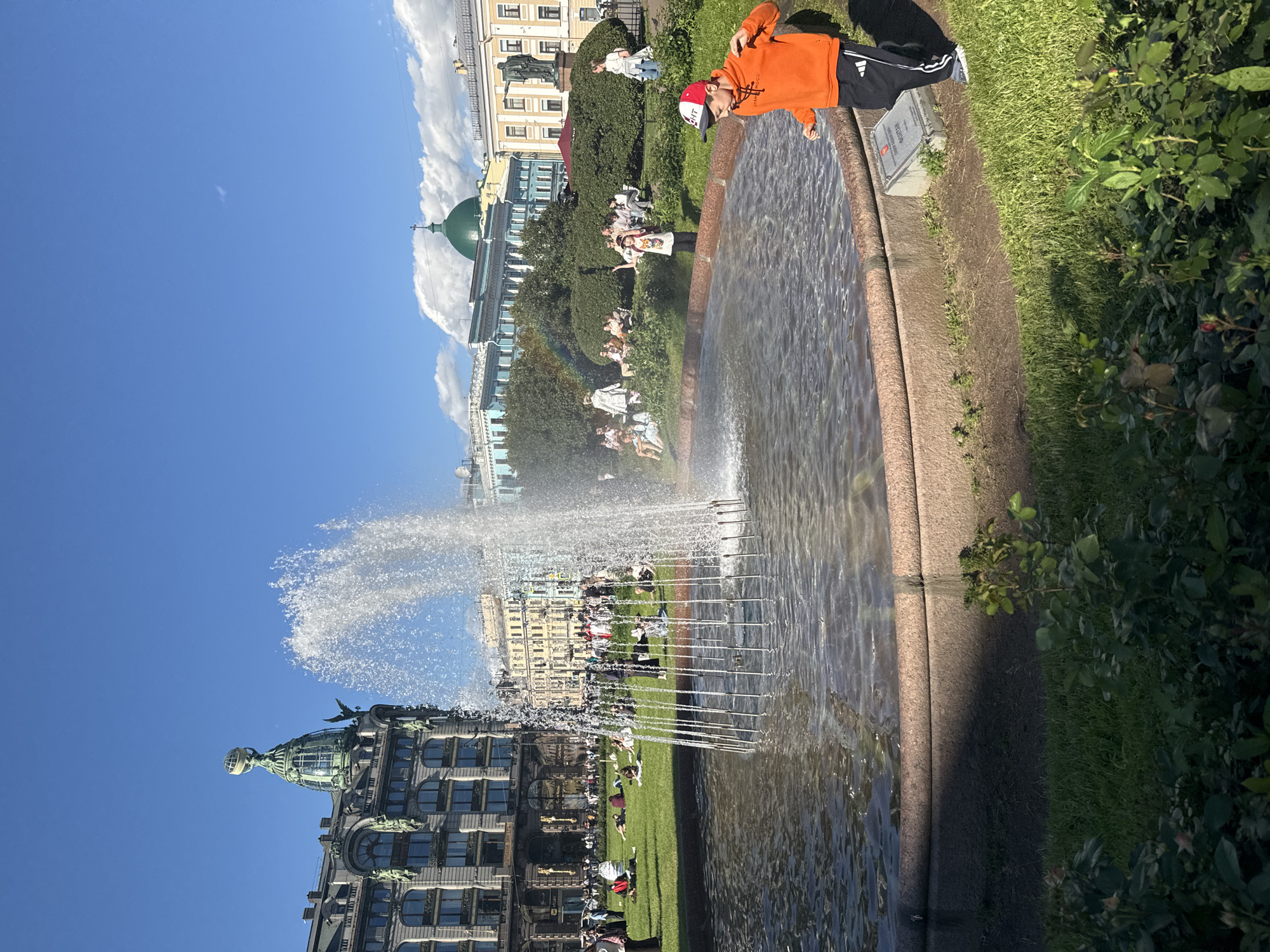 The image depicts a sunny day in Palace Square, Saint Petersburg, Russia. In the foreground, a young boy in an orange hoodie and a baseball cap stands near a large, circular fountain with water spraying upwards. The boy is wearing black pants with white stripes. Behind the fountain, a lush green lawn is dotted with people relaxing and sitting. In the midground, a group of people are gathered in the shade of trees and bushes. The iconic Singer House, with its distinctive globe-topped dome, stands prominently to the left of the frame. Several other historical buildings with ornate facades and domed roofs, characteristic of Saint Petersburg's architecture, are visible in the background under a bright blue sky with scattered clouds. A faint rainbow is visible in the mist of the fountain.