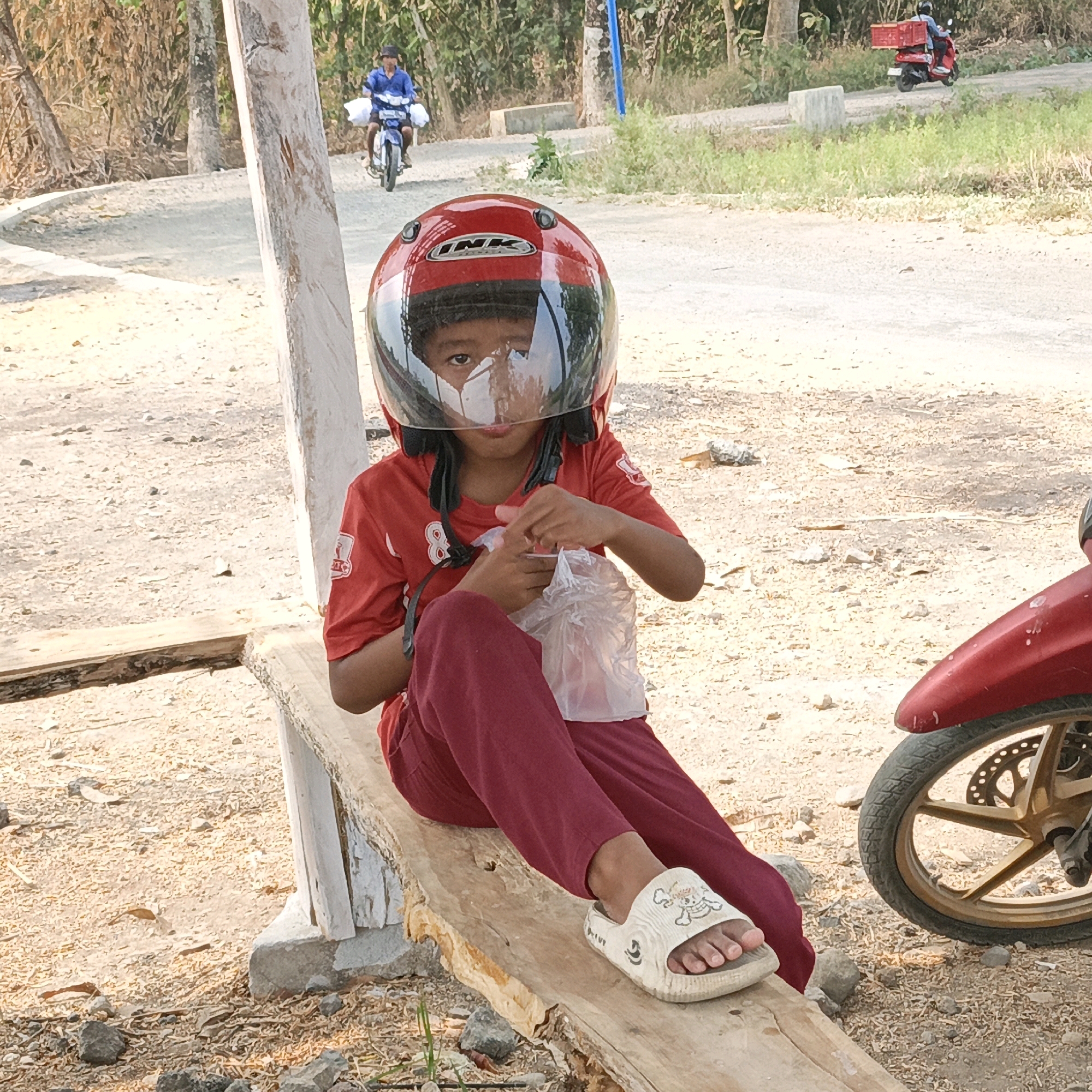 In Purana, Indonesia, a child wearing a red motorcycle helmet and a red shirt sits on a wooden bench. The child is holding a clear plastic bag. The child's face is partially obscured by the helmet's visor and a white mask. Maroon pants and white sandals with cartoon characters complete the child's attire. To the right of the child, the front wheel and fender of a red motorcycle are visible. The background features a dirt road, trees, and foliage. In the distance, two other individuals on motorcycles can be seen. One motorcycle has a red crate attached to the back. The scene appears to be outdoors in daylight.