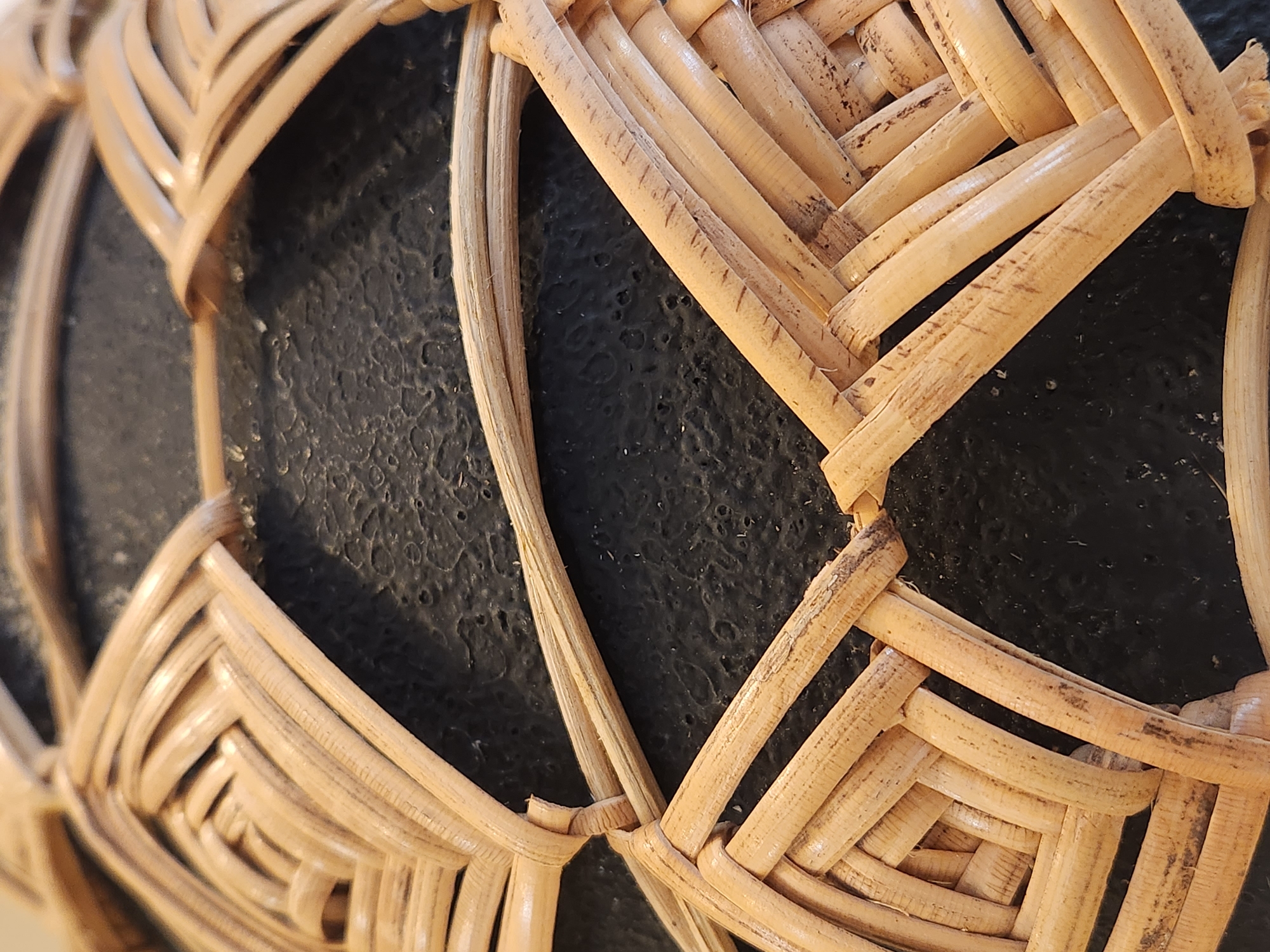 The image is a close-up, macro shot of a woven wicker material, likely a basket or decorative item, against a dark, textured surface. The wicker is made of natural, light-colored reeds or bamboo, showcasing a detailed woven pattern with some natural imperfections and darker markings on the material. The dark background has a bumpy, irregular texture, resembling a rough stone or painted surface. The lighting is direct, highlighting the texture of both the wicker and the background. There are no people or animals present in the image. The location in Maitland, United States of America, is not identifiable from the visual information provided.
