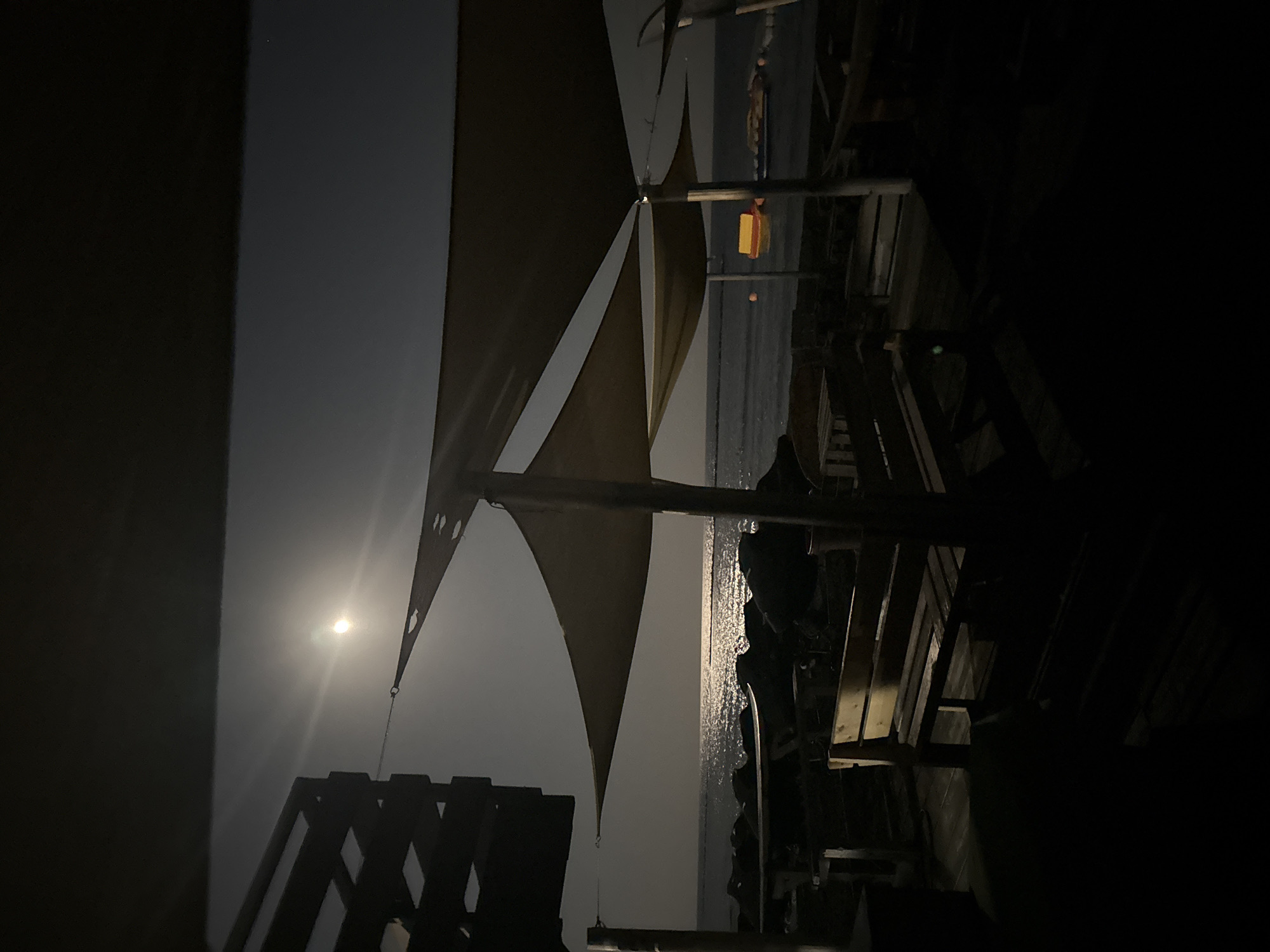 The image depicts a nighttime scene at a beachside establishment in Agios Tychona Community, Cyprus. The moon is visible and shining brightly, casting a shimmering reflection on the calm sea. Several canvas shade structures are erected over an outdoor seating area. Wooden benches and tables are present, some of which are illuminated by the moonlight. In the water, there are some inflatable water toys. The foreground is largely in shadow, obscuring details of the immediate seating area. The overall atmosphere is tranquil and serene.