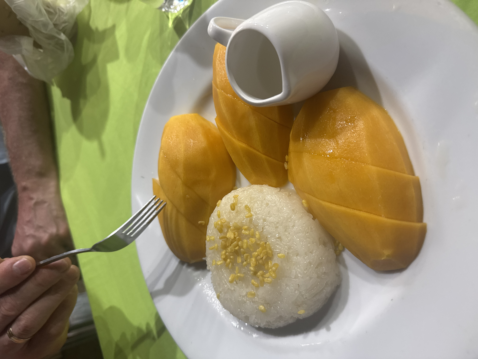 A close-up shot depicts a plate of mango sticky rice, a popular Thai dessert, likely being consumed. The plate contains sections of ripe yellow mango, a mound of sticky rice sprinkled with yellow crispy fried mung beans, and a small white pitcher, presumably containing coconut milk sauce to pour over the dish. A hand holding a fork is visible, indicating that someone is about to eat the dessert. The setting appears to be a restaurant or dining table, with a green surface visible in the background. While the city of Patong, Thailand, is mentioned, there is no visual element within the image to confirm this location. The image focuses primarily on the dessert itself and the immediate action of eating it.