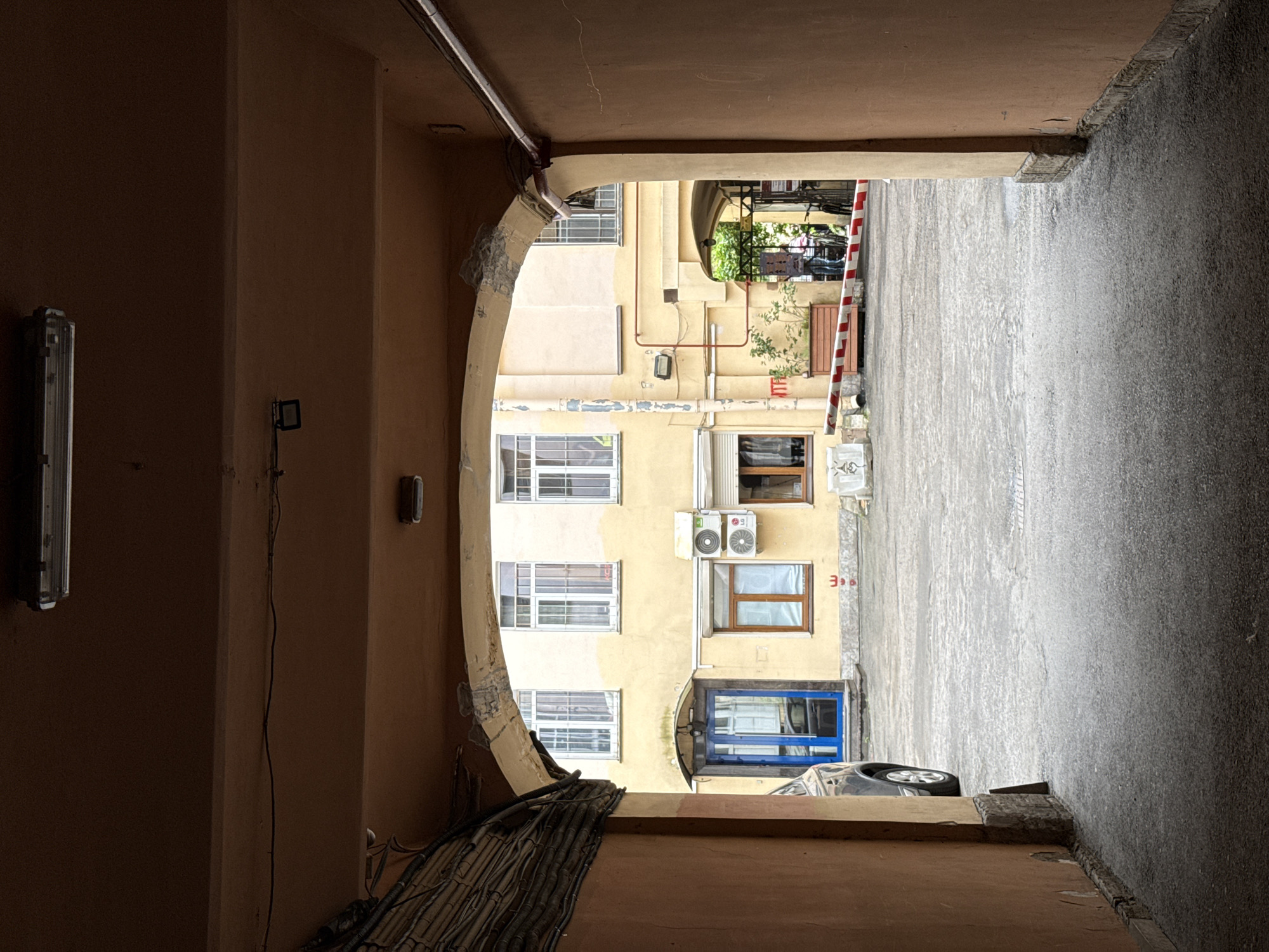 This is an eye-level, medium shot captured from within a dark, arched passageway looking out into a courtyard in Saint Petersburg, Russia. The courtyard is paved with concrete and shows signs of wear and tear. A yellow-colored building with white-framed windows dominates the background.
To the left, a dark-colored car is parked partially in the frame, with its front end obscured. Adjacent to the car is a vibrant blue door. Further into the courtyard, an air conditioning unit is mounted on the building's facade. To the right, a red and white striped barrier is partially visible, suggesting an entry or exit point. In the distance, behind the barrier, two bicycles and two individuals can be seen, one of whom appears to be interacting with a dog, though details are indistinct.
The passageway itself is characterized by its warm, orange-hued walls and a rough, textured ceiling. Electrical conduits and wiring are visible along the left side of the passageway ceiling, and a fluorescent light fixture is mounted overhead. The overall impression is one of a utilitarian, somewhat aged urban environment.