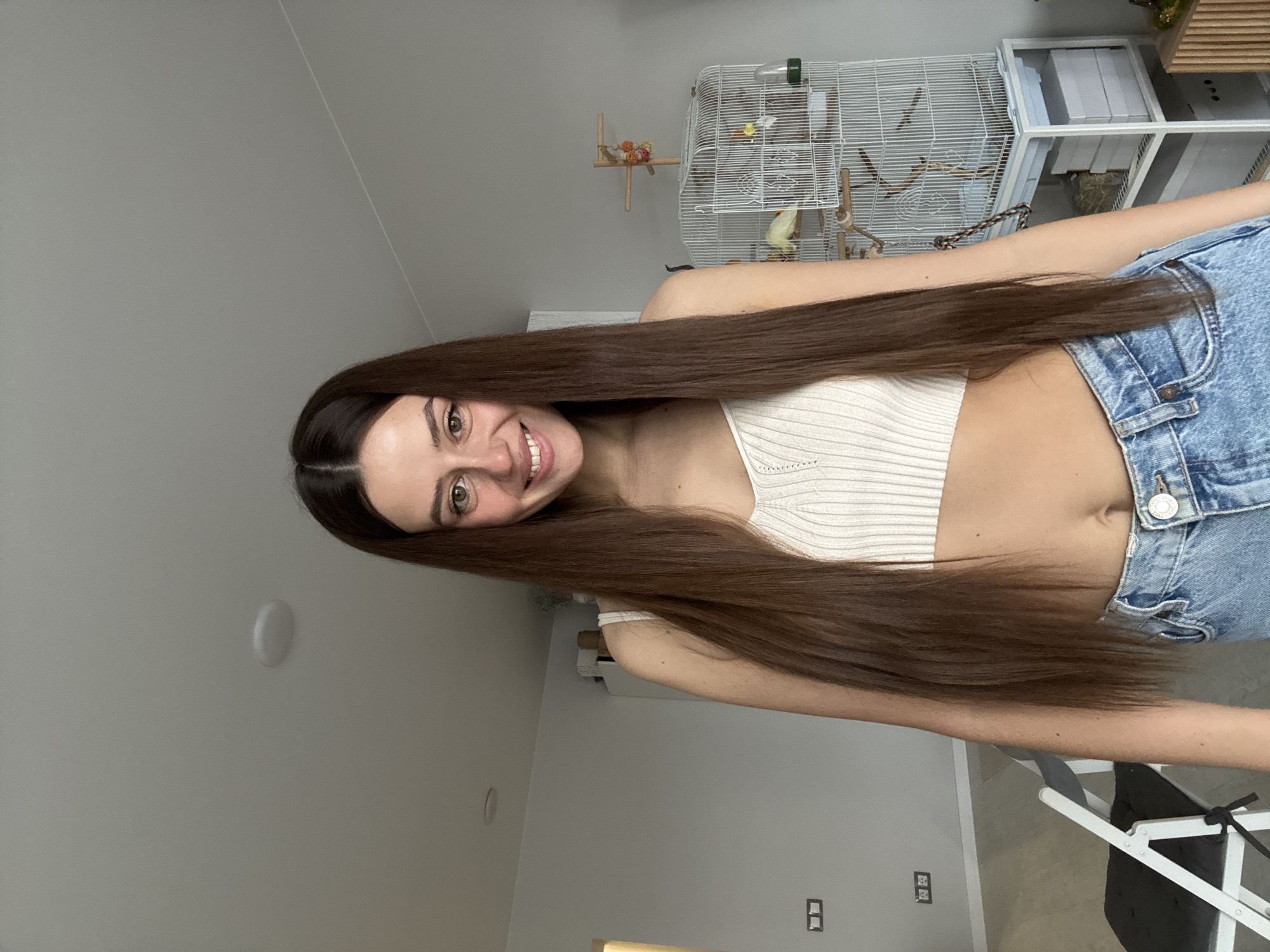 A young woman with long, straight brown hair is smiling at the camera. She is wearing a light-colored knit crop top and light blue denim jeans. In the background, there is a white birdcage with a small yellow bird inside. A white shelf unit with items on it is next to the birdcage. The walls are painted in a light neutral color. There are electrical outlets visible on the wall. The image appears to be taken indoors, with natural light. There is not enough information to determine the location in Krasnoyarsk, Russia.