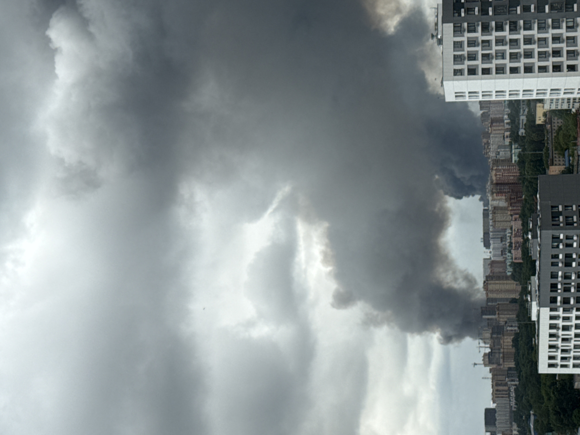 A panoramic view captures a city skyline under a cloudy sky, with thick plumes of dark smoke rising from a densely populated area. The smoke appears to originate from a section of buildings in the mid-ground, obscuring the view of the cityscape behind it. In the foreground, modern residential buildings with numerous windows are visible, one prominently featuring a white facade with gray accents. The sky is overcast with heavy, gray clouds. The image is likely taken from an elevated position, offering a broad perspective of the urban environment. This scene is identified as being in Lyubertsy, Russia.
