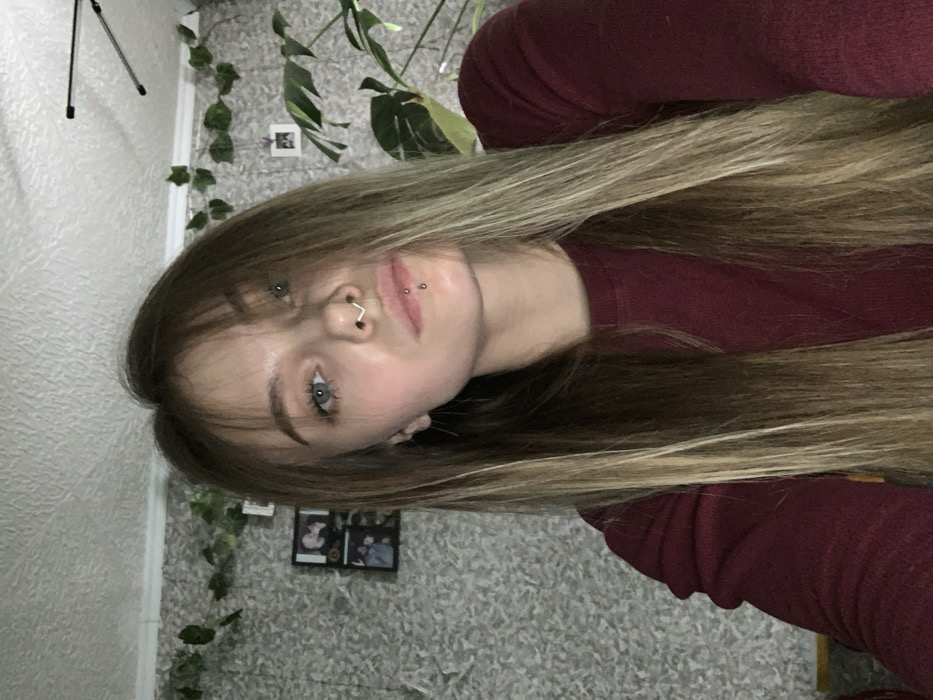 An indoor portrait features a young female subject. She has long, straight, two-toned hair with bangs, light blue eyes, and fair skin. The subject displays a silver-toned V-shaped septum piercing and a vertical labret piercing on her lower lip. She wears a maroon long-sleeved crew-neck top and looks directly into the camera with a neutral expression.
The background reveals a room with light gray or white textured wallpaper. Artificial green ivy is draped along the upper portion of the wall. Two framed photographs are visible on the left wall, and a smaller white-framed photo hangs further to the right. A large green houseplant is situated behind the subject on the right side of the frame. A black tripod or stand is partially visible in the upper right corner against a textured white ceiling.
The specific city of Фоки, Russia, cannot be determined from the visual information present in the image. The scene is consistent with a residential or private indoor setting.