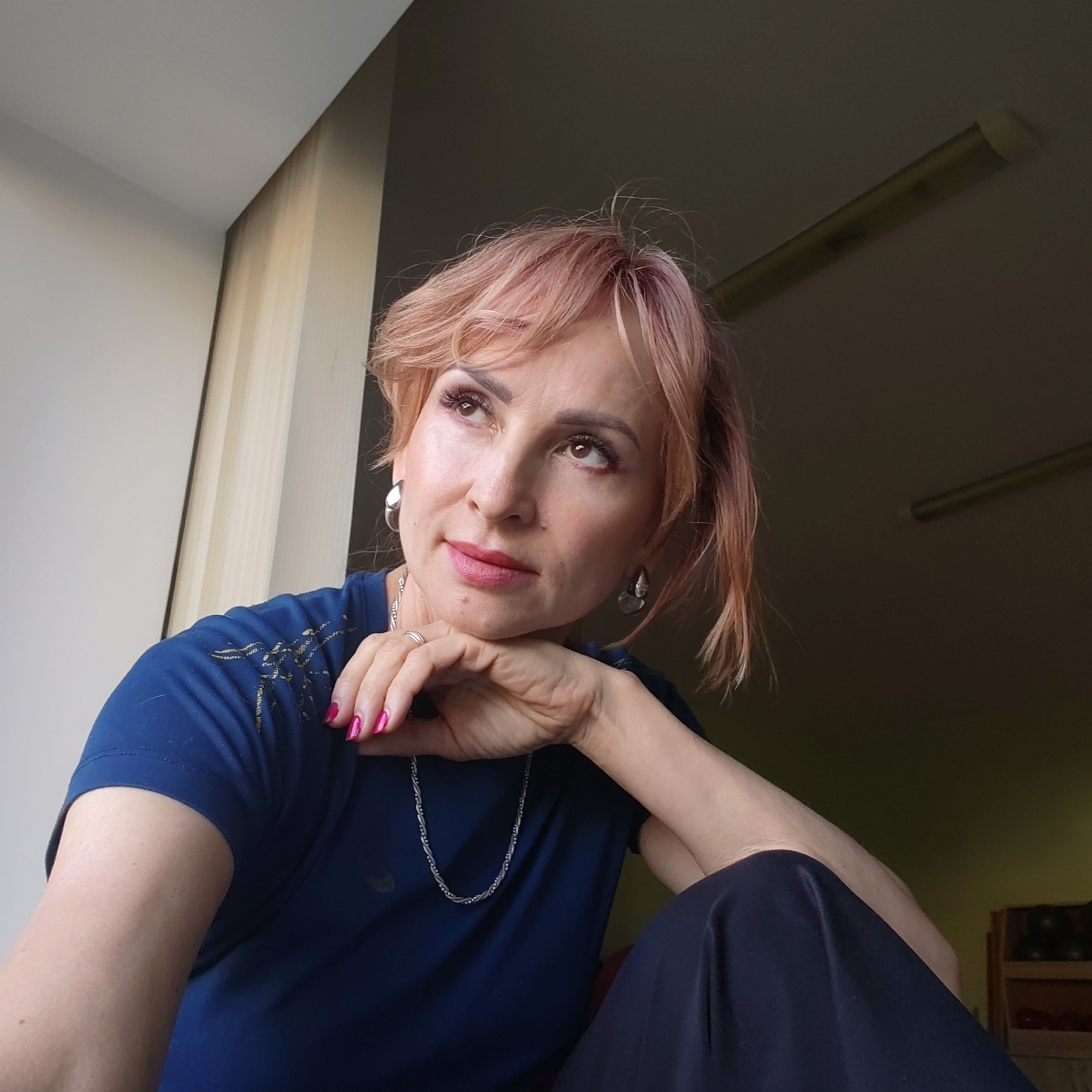 A fair-skinned woman with pinkish-blonde hair is the focus of the image. She wears a dark blue t-shirt with some light-colored design on the shoulder and a silver necklace, also earrings. Her nails are painted a bright pink. She rests her chin on her hand and looks upward and to the left. The background is a muted interior, with a window on the left and a light fixture on the ceiling on the right. There is furniture visible in the background as well.
The location is identified as the city of Chaikovsky, Russia, though there is no feature in the image that suggests this definitively.