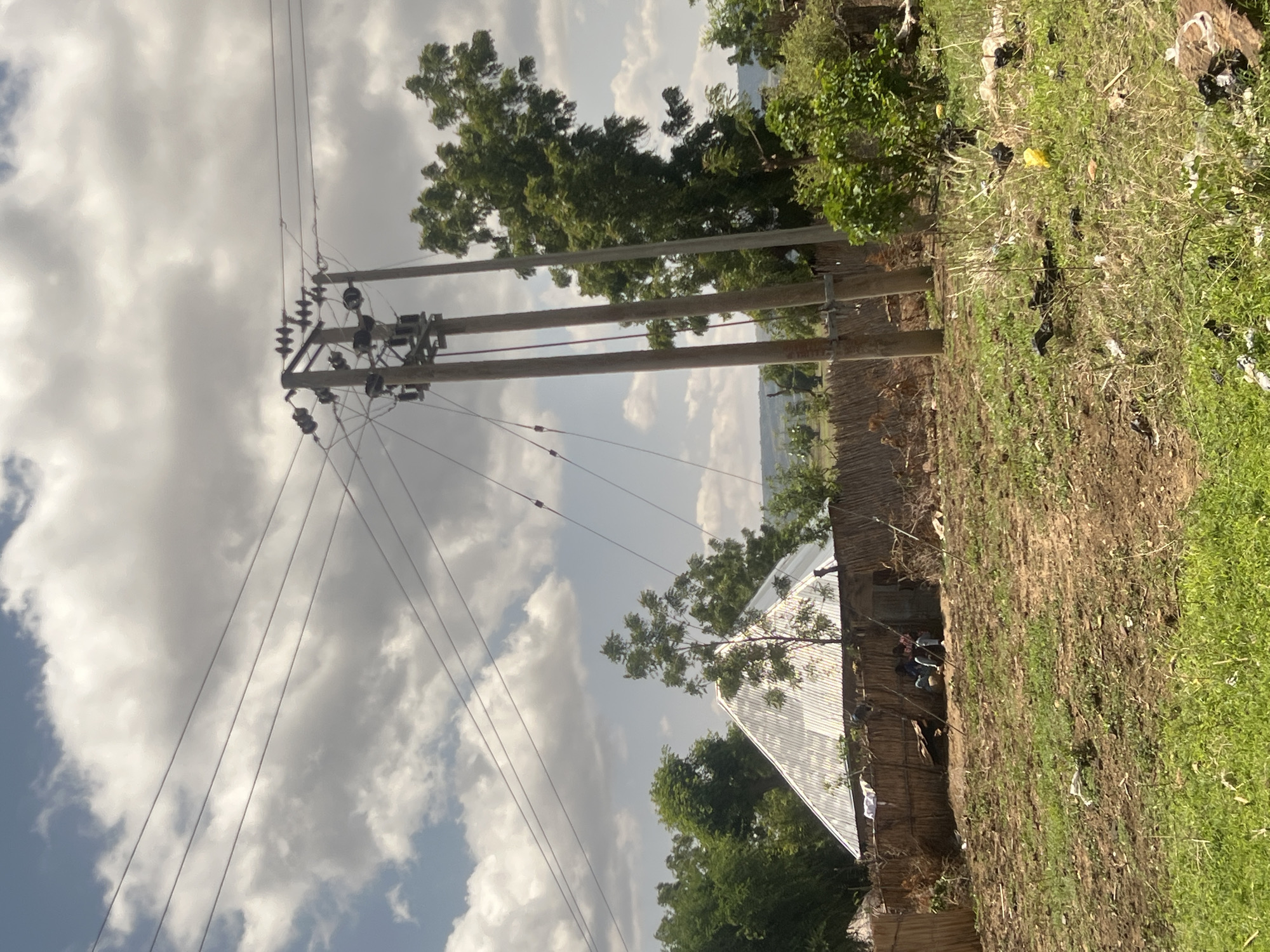 This image depicts a semi-rural outdoor scene in the city of Chum, Nigeria, under a sky with prominent white cumulus clouds against a light blue background.
In the foreground, the ground consists of uneven, reddish-brown soil mixed with sparse green grass and some dry vegetation. Scattered litter, including white plastic bags, dark plastic debris, and at least one yellow plastic item, is visible across the lower half of the frame.
The mid-ground features a cluster of three tall, weathered wooden utility poles positioned vertically from the lower right to the upper right. These poles are equipped with multiple cross-arms supporting ceramic or glass insulators and a complex arrangement of overhead electrical wires. A distinct red cable runs vertically down one of the central poles. To the left of these poles, a structure with a reflective, corrugated metal pyramidal roof is visible, partially obscured by green trees. Below and in front of this structure is a low fence constructed from woven dark brown organic material, possibly reeds or sticks, extending horizontally across the frame.
Two individuals are observed in the mid-left, sitting or crouching on the ground within an opening behind the woven fence, near the base of the pyramidal roof structure. They are wearing dark clothing, including blue jeans, and appear to be at rest.
Numerous green trees and bushes are scattered throughout the scene, providing foliage behind the fence, around the structures, and in the background. A distinct, slender tree stands to the left of the main utility poles. The background reveals a hazy distant landscape with further vegetation and what appears to be a low-lying horizon.