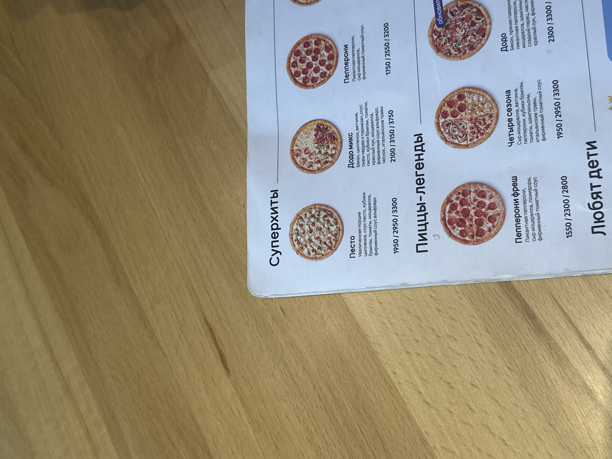 The image shows a menu page from a restaurant, likely a pizzeria, with various pizza options listed. The menu is printed on white paper with black text and features colorful images of pizzas. The pizzas are arranged in rows, with prices listed below each. The text is in Russian. The menu is lying on a wooden surface. There are no people or other objects visible in the image. The location of this restaurant in Gyumri, Armenia, cannot be determined from the image alone.