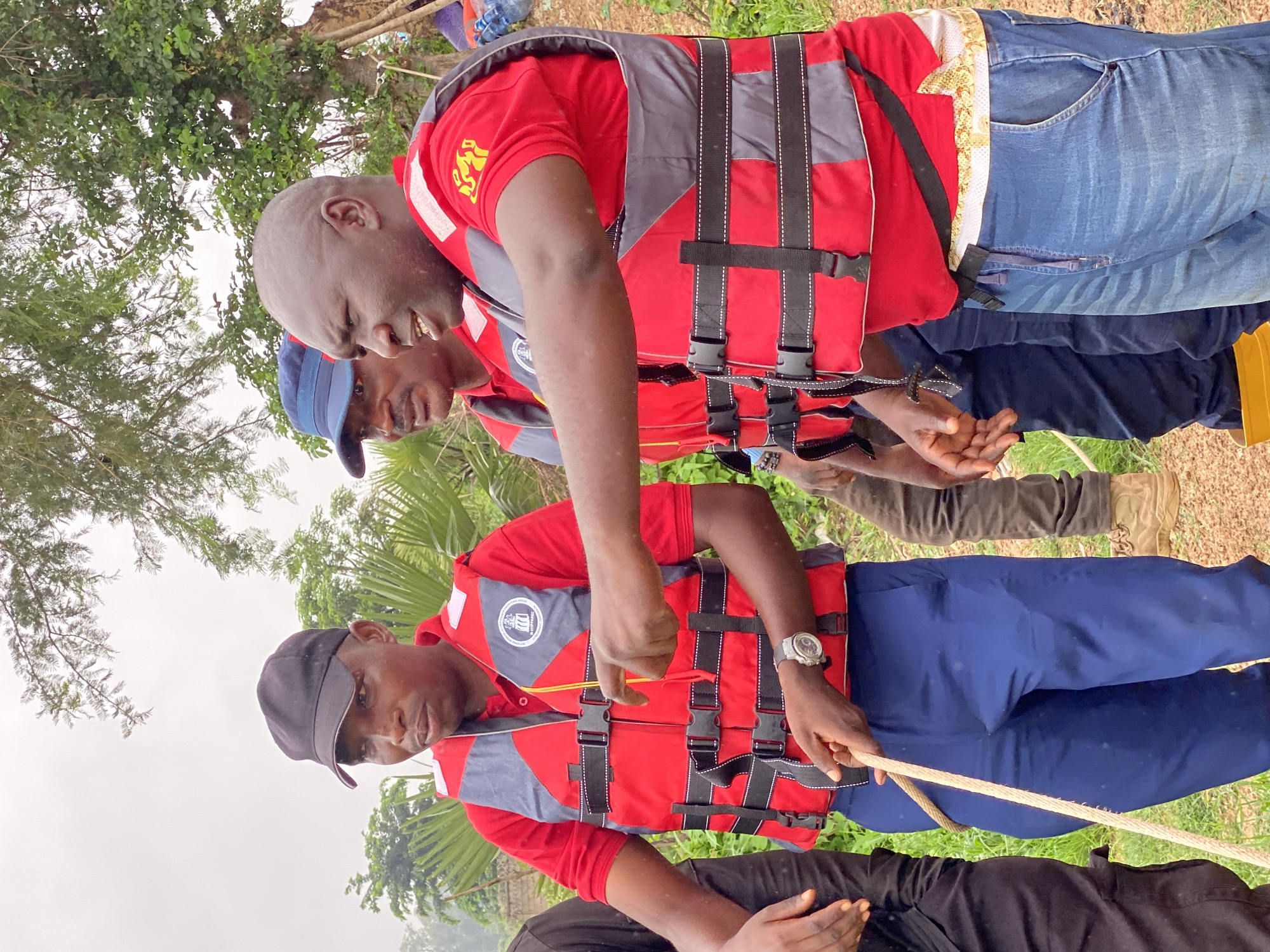 Three men are standing outdoors in what appears to be a rural or semi-rural area. The man in the foreground, wearing a red life vest over a red shirt and blue pants, is holding a thick rope. He has a black baseball cap on and a watch on his left wrist. To his right, another man, wearing a blue cap and a similar red life vest over a red shirt, is looking towards the right. A third man, partially visible behind them and to the left, is wearing a red life vest over a black shirt. The background shows lush green vegetation, including palm-like plants and trees, with an overcast sky. There is a dirt path or ground visible in the lower right portion of the image. The presence of life vests and the rope suggests a context related to water activities or rescue operations. The image is likely taken in Kawo, Nigeria, given the context provided.