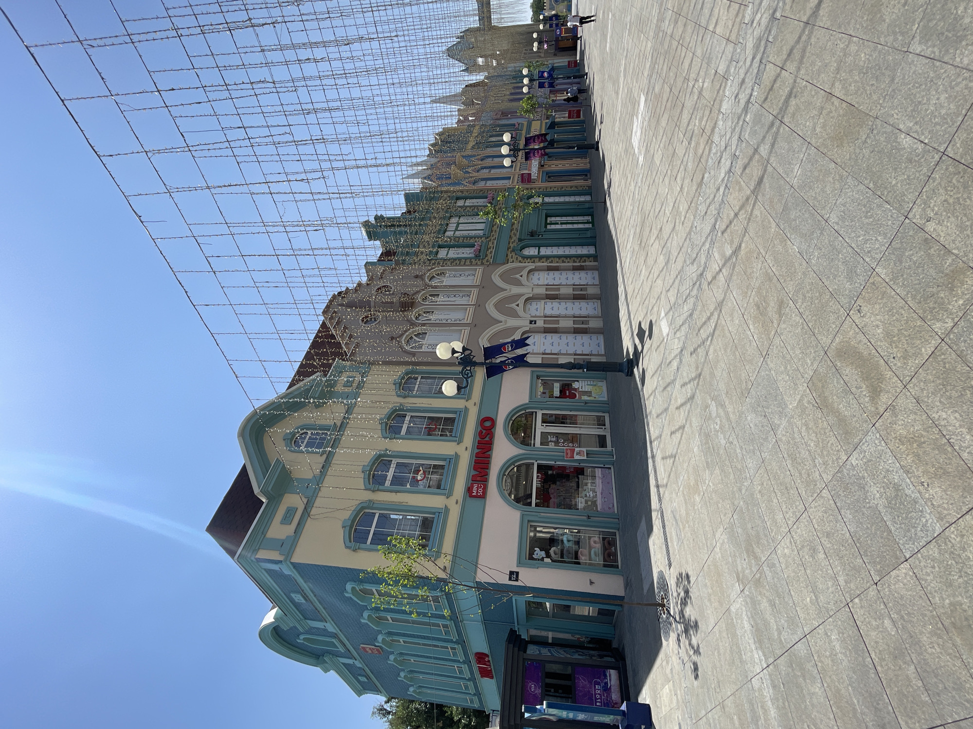 This media file shows a street scene with buildings and a pedestrian walkway. The buildings have an ornate, old-world architectural style with pastel-colored facades, arched windows, and decorative trim. They are adorned with strings of small, white lights that hang overhead, creating a festive atmosphere. One building prominently displays the "MINISO" logo in red lettering. The walkway is paved with large, grey tiles, and a tree with sparse foliage casts a shadow on the ground. The sky is a clear, bright blue, indicating a sunny day. The perspective suggests the photo was taken looking upwards and slightly to the right from ground level. A few people are visible in the distance, walking along the street. The overall impression is of a well-maintained, possibly tourist-oriented street in a city, with a cheerful and bright ambiance. The location context of Tashkent, Uzbekistan, suggests this could be a popular shopping or entertainment district. There is also text visible on some banners and signage, including "MINISO" and what appears to be "BLOOM" on a dark blue sign at the bottom of the frame.