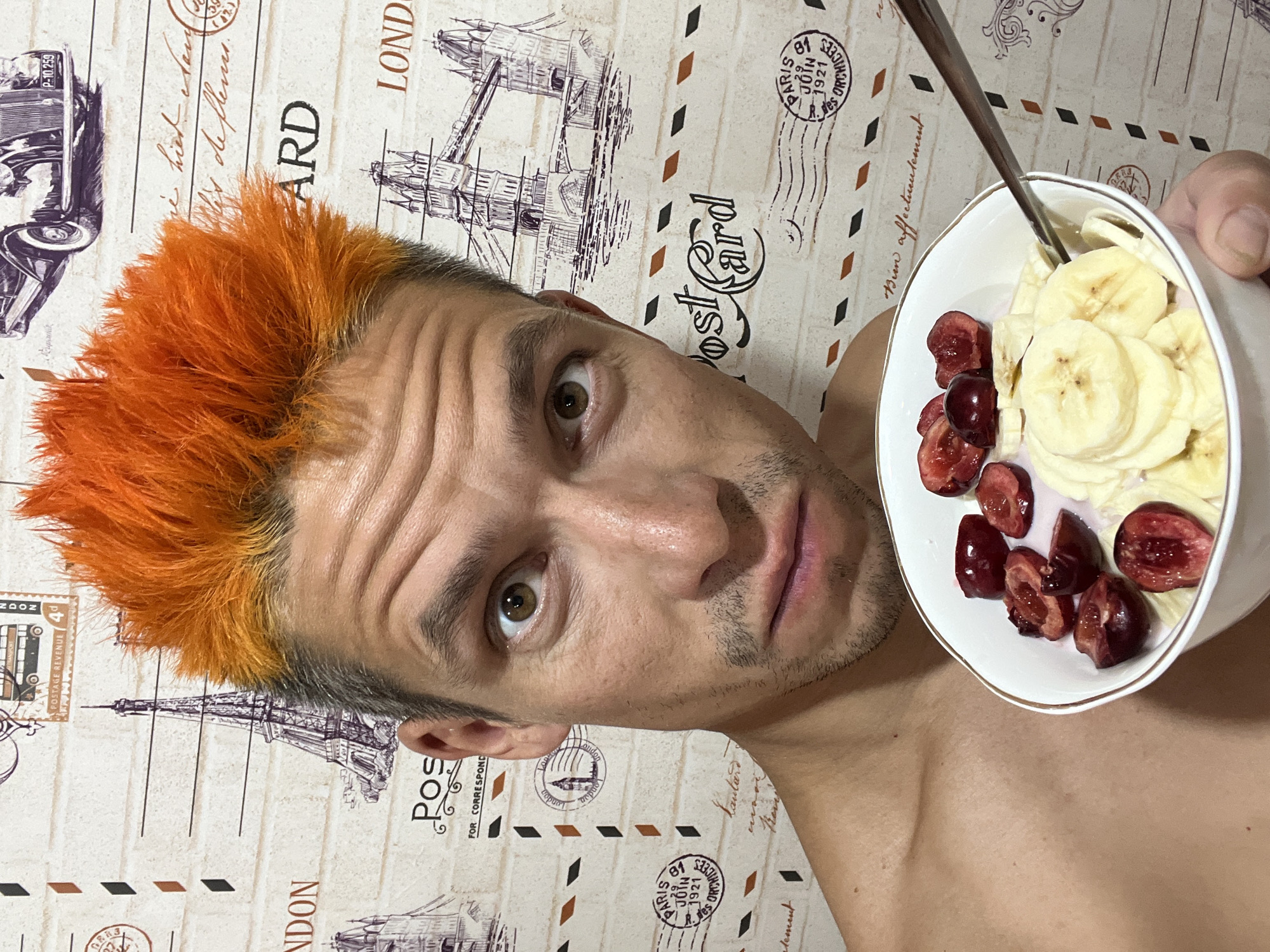 A man with bright orange hair and stubble on his face is pictured holding a white bowl filled with sliced bananas and halved red cherries or berries, with a spoon resting in it. He is looking directly at the camera with a neutral to slightly surprised expression, his bare chest visible. The image is taken at an angle, as if a selfie.
The background features a light-colored surface, possibly wallpaper or fabric, adorned with a vintage postcard and travel theme. Visible illustrations include a classic car, the Eiffel Tower, and London Bridge. Various texts and stamps are printed on this background. Notable text includes "LONDON", "POST CARD", "PARIS JOIN 1921", "POSTAGE REVENUE", "P-10.259", "Bien affectueusement", "Pos FOR CORRESPOND", and other partially visible words in what appears to be French. The scene is indoors, and the time of day is not discernible. The provided location context is Chaikovsky, Russia.