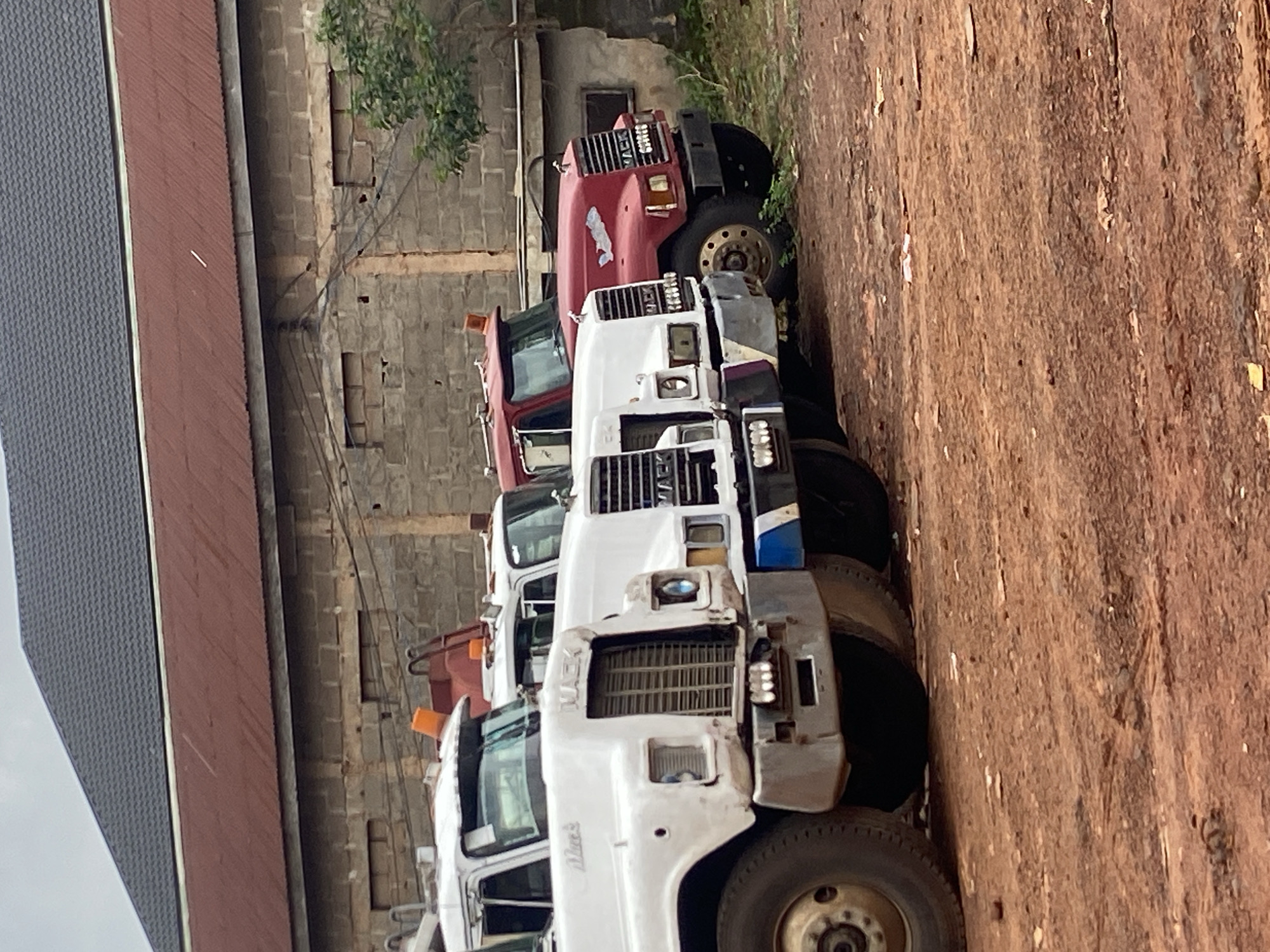 In Nkpor, Nigeria, a row of parked Mack trucks is lined up in front of a brick building with a corrugated metal roof. The trucks, in various states of repair, are positioned on a dirt lot. The foreground is dominated by the trucks, with the white truck on the left being the most prominent, followed by another white truck, and then a red truck on the right. The building behind them appears to be a workshop or storage facility. The sky is overcast, suggesting a cloudy day.