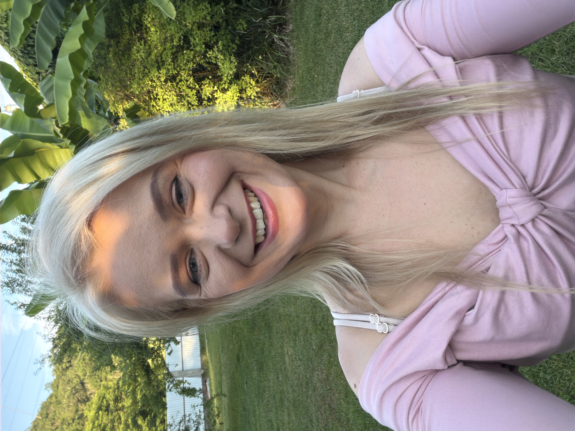 A young woman with blonde hair is pictured in a selfie outdoors. She is wearing a pink off-the-shoulder top that is tied at the bust. Her hair is long and straight, and some of it falls across her face. Her makeup includes eyeshadow and lipstick. She is smiling, showing her teeth. The background of the image consists of lush green grass and trees, suggesting a natural outdoor setting. There is also a hint of a white fence or structure in the background, possibly indicating the edge of a property or park. The lighting suggests it is daytime, likely late afternoon or early evening, given the warm glow on her face and the shadows. The overall mood appears relaxed and happy. The image is oriented vertically.