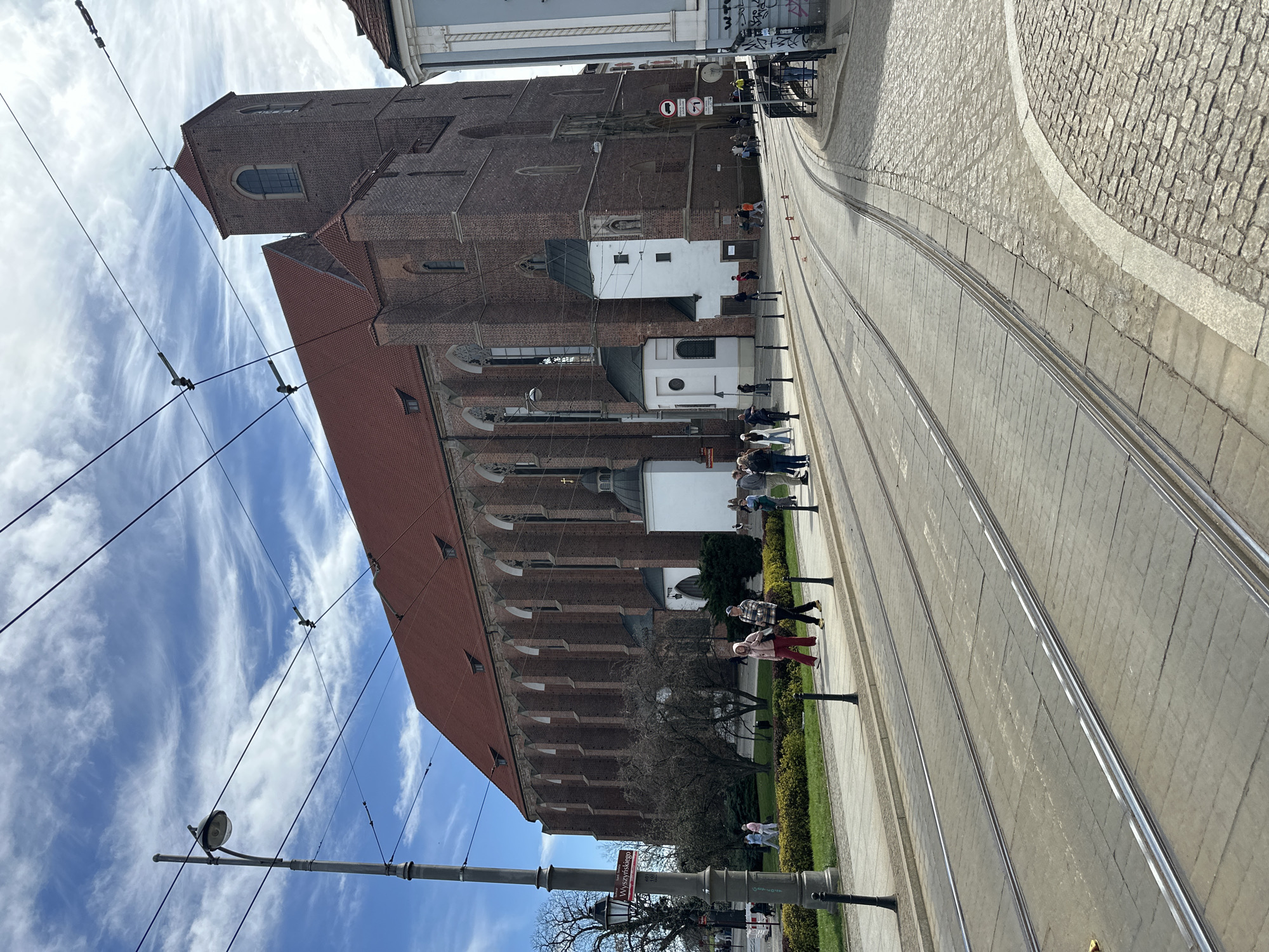 The image shows the Collegiate Church of the Holy Cross and St. Bartholomew in Wrocław, Poland, from street level. A tram line runs parallel to the church along the street. Several pedestrians are walking alongside the church. The sky is visible above the church and contains numerous clouds. There are utility poles and wires near the edge of the frame. A road sign can be seen near a tree, towards the bottom of the frame. The roadway next to the tram tracks is made of cobblestone.