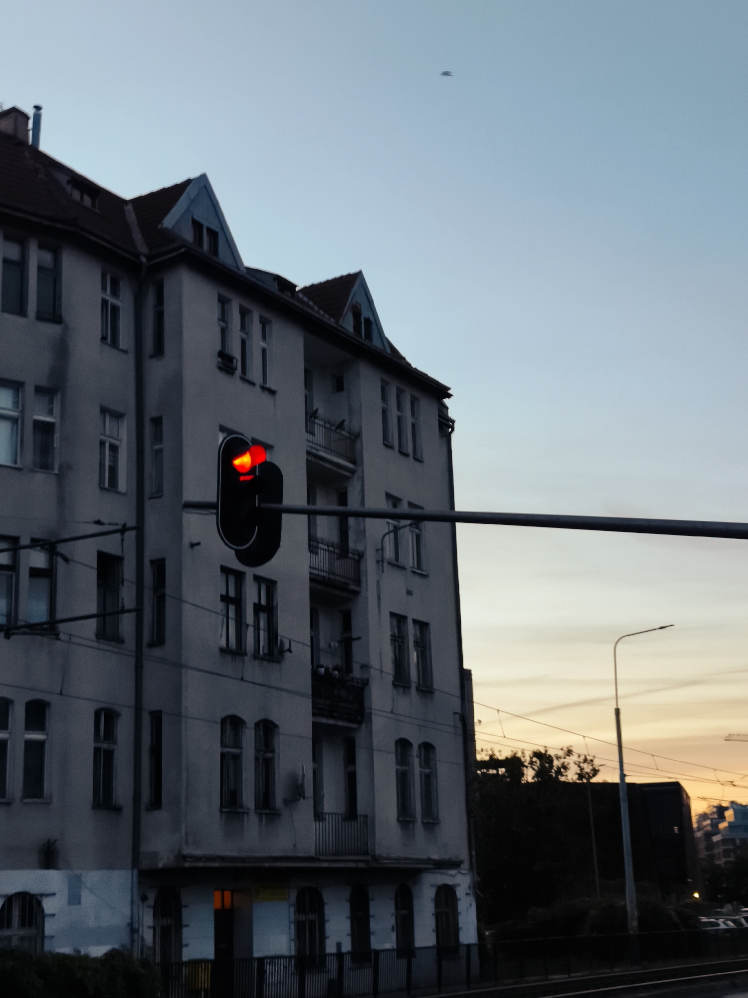 The image depicts a street scene at dusk in the city of Gdańsk, Poland. The focal point is a multi-story apartment building, likely residential, with a grey facade and a dark red tiled roof. Dormer windows are visible on the top floor. In the foreground, a traffic light displays a red signal, indicating a stop. Overhead wires, possibly for an electric tram system, are suspended across the scene. In the background, silhouetted trees and a street lamp suggest an urban environment. The sky is a gradient of pale blue and orange, indicative of sunset. A faint, dark, elongated shape is visible in the upper sky, its nature unclear. The scene appears devoid of people.