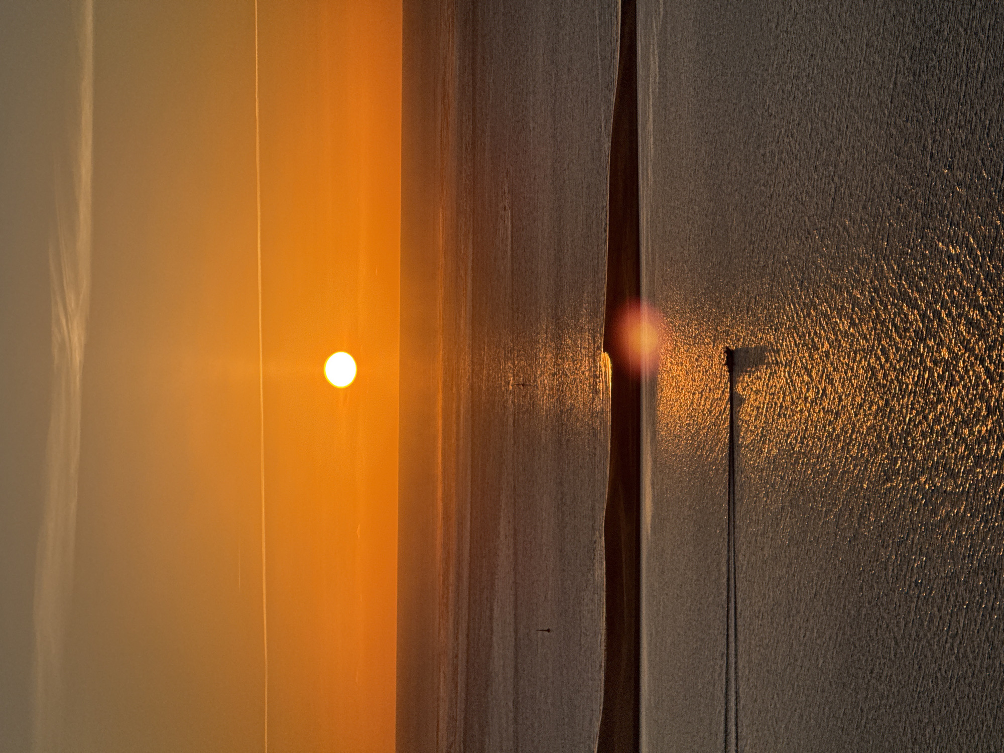 The image depicts a serene sunset over the Arcachon Bay in La Teste-de-Buch, France. The sun, a bright orb, hangs low on the horizon, casting a warm, orange glow across the sky and water. A thin contrail stretches across the upper portion of the sky, a subtle indication of human activity.
The water itself is a gradient of deep blues and grays, reflecting the fiery hues of the sunset. A long, narrow strip of land, likely the Dune of Pilat, forms a dark silhouette across the middle ground, separating the bay into two distinct bodies of water.
In the foreground, a small boat is making its way across the water, leaving a subtle wake behind it. The boat is silhouetted against the shimmering reflection of the sun on the water, creating a dynamic contrast. The scene is one of tranquility and natural beauty, capturing the essence of a peaceful evening by the coast.