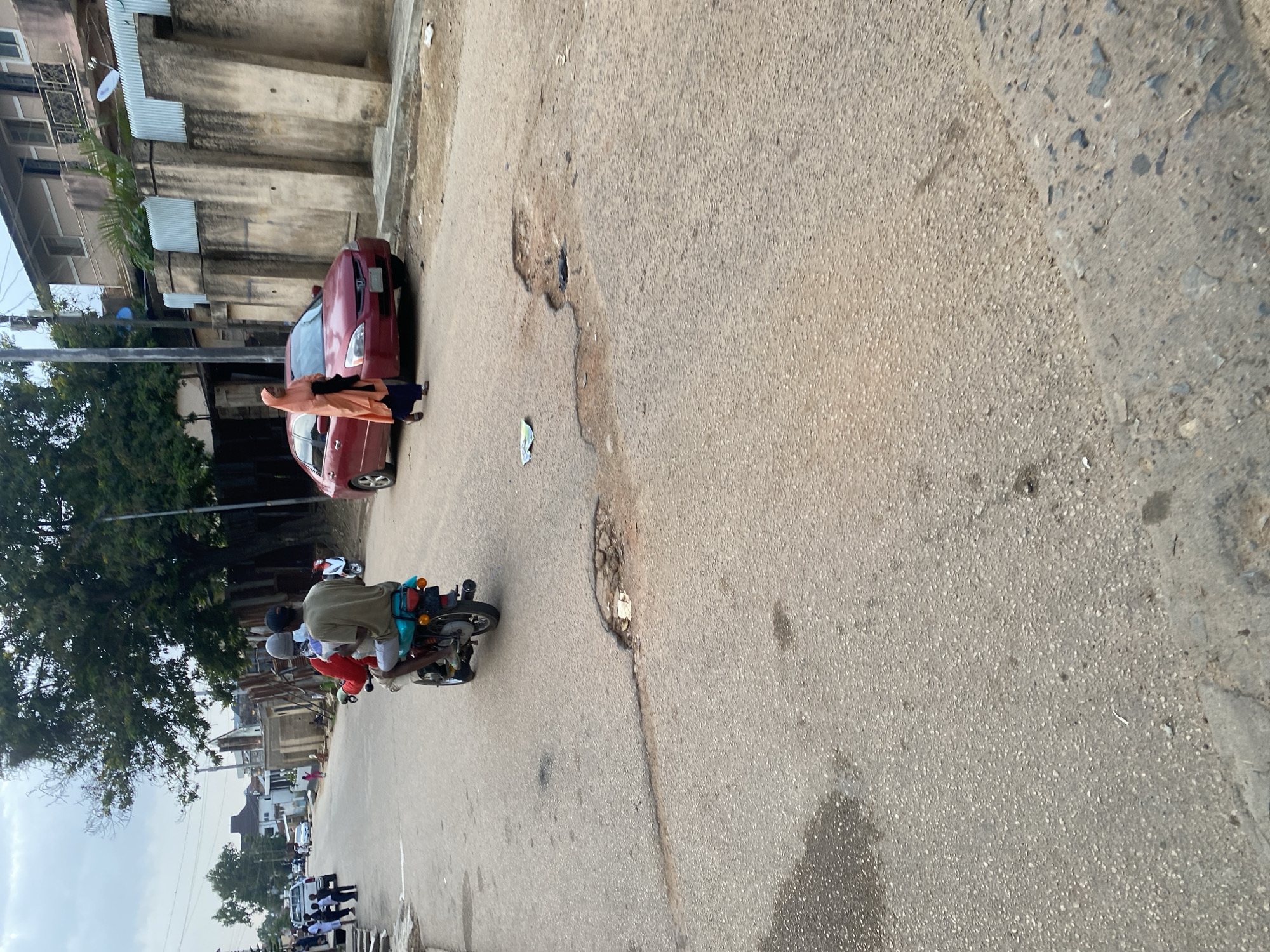 The image depicts a street scene in Jos, Nigeria, characterized by a worn asphalt road with several visible potholes. In the foreground, a motorcycle with two male occupants is moving away from the camera. The rider wears a light-colored t-shirt and a head covering, while the passenger in front wears a red shirt and a dark head covering. The motorcycle is a light blue color. Further down the road, a red sedan is parked. A woman wearing an orange hijab and dark trousers walks in the opposite direction of the motorcycle, passing the parked car. Buildings with concrete walls and corrugated metal fencing line the right side of the street, with trees and overhead utility lines visible in the background. The sky is overcast.